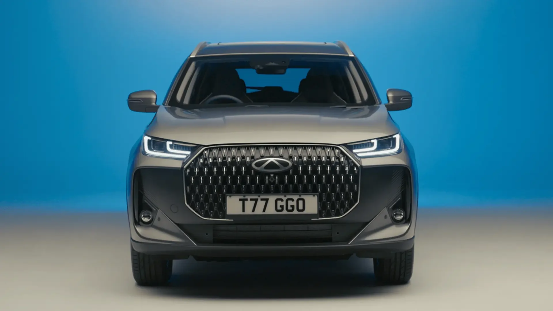 Front view of a modern gray SUV with a large grille, sleek headlights, and a license plate reading T77 GGO, against a blue background.