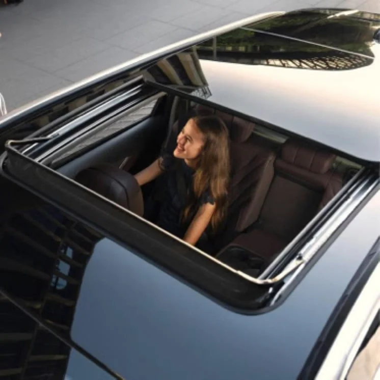 A woman smiling and sitting in the backseat of a black car with an open sunroof, taken from above.