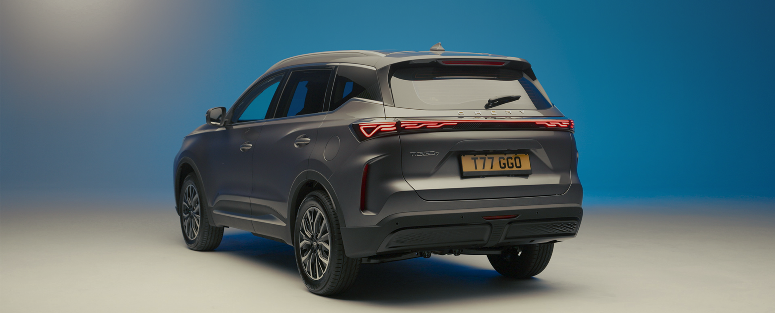 Rear view of a matte gray SUV, a Chery Tiggo 7, on a plain background with a blue gradient, showcasing its taillights, license plate, and design details.