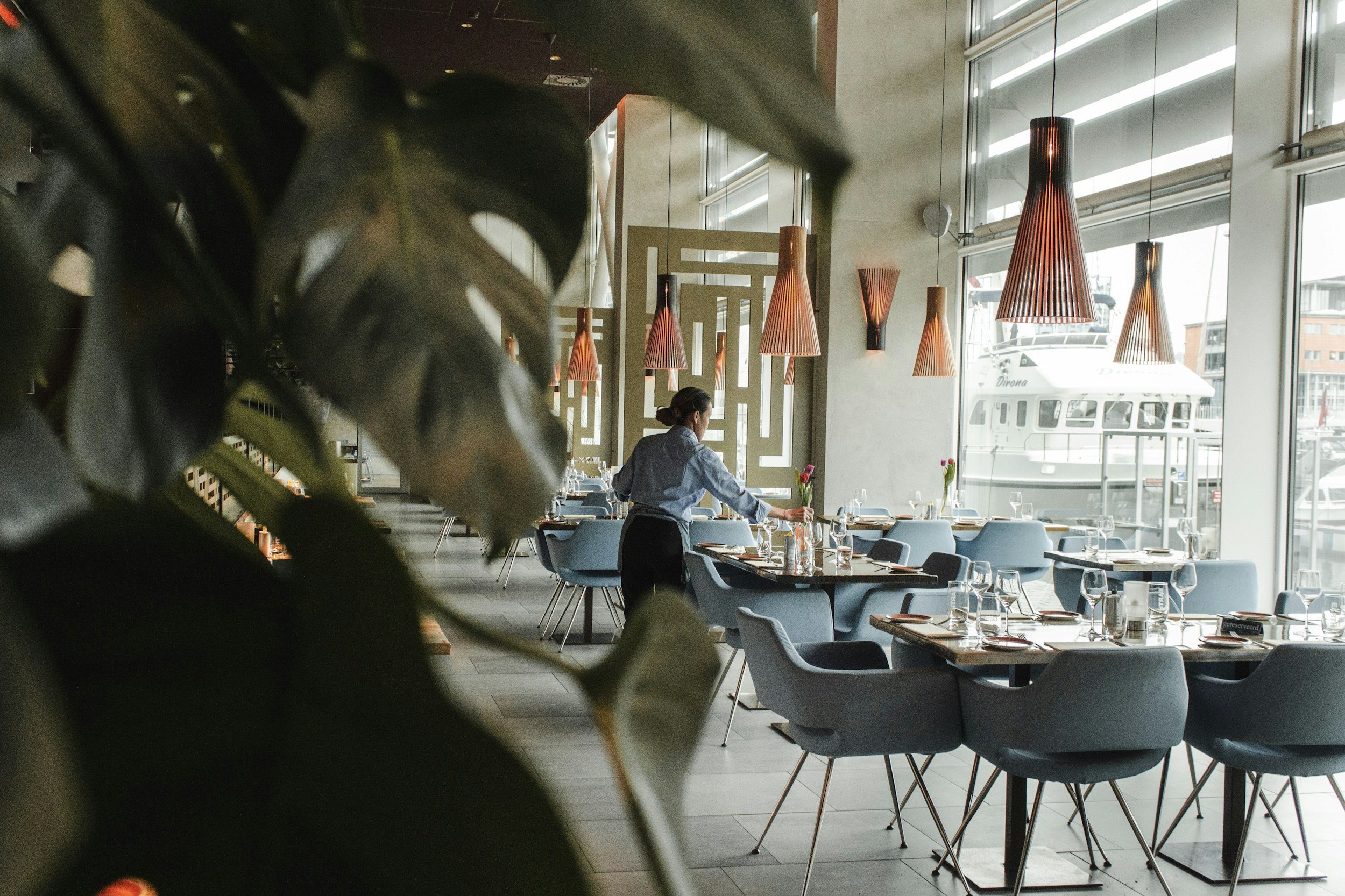 A modern restaurant with large windows, wooden pendant lights, and blue chairs. A waitress is setting a table with glasses and a vase with pink flowers, with a view of a marina outside.