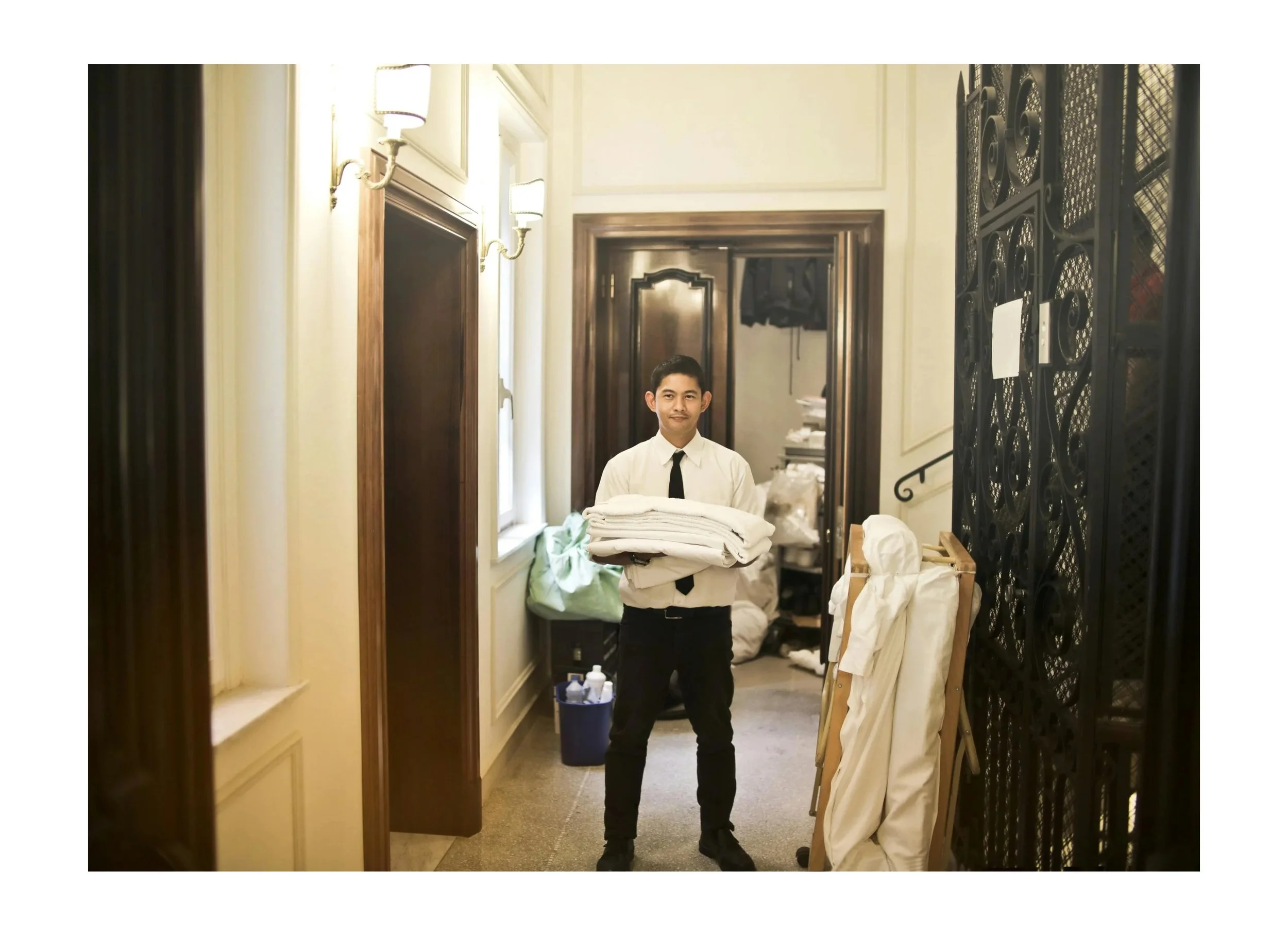 A male hotel staff member in a white shirt and black tie holding folded linens in a hotel corridor.