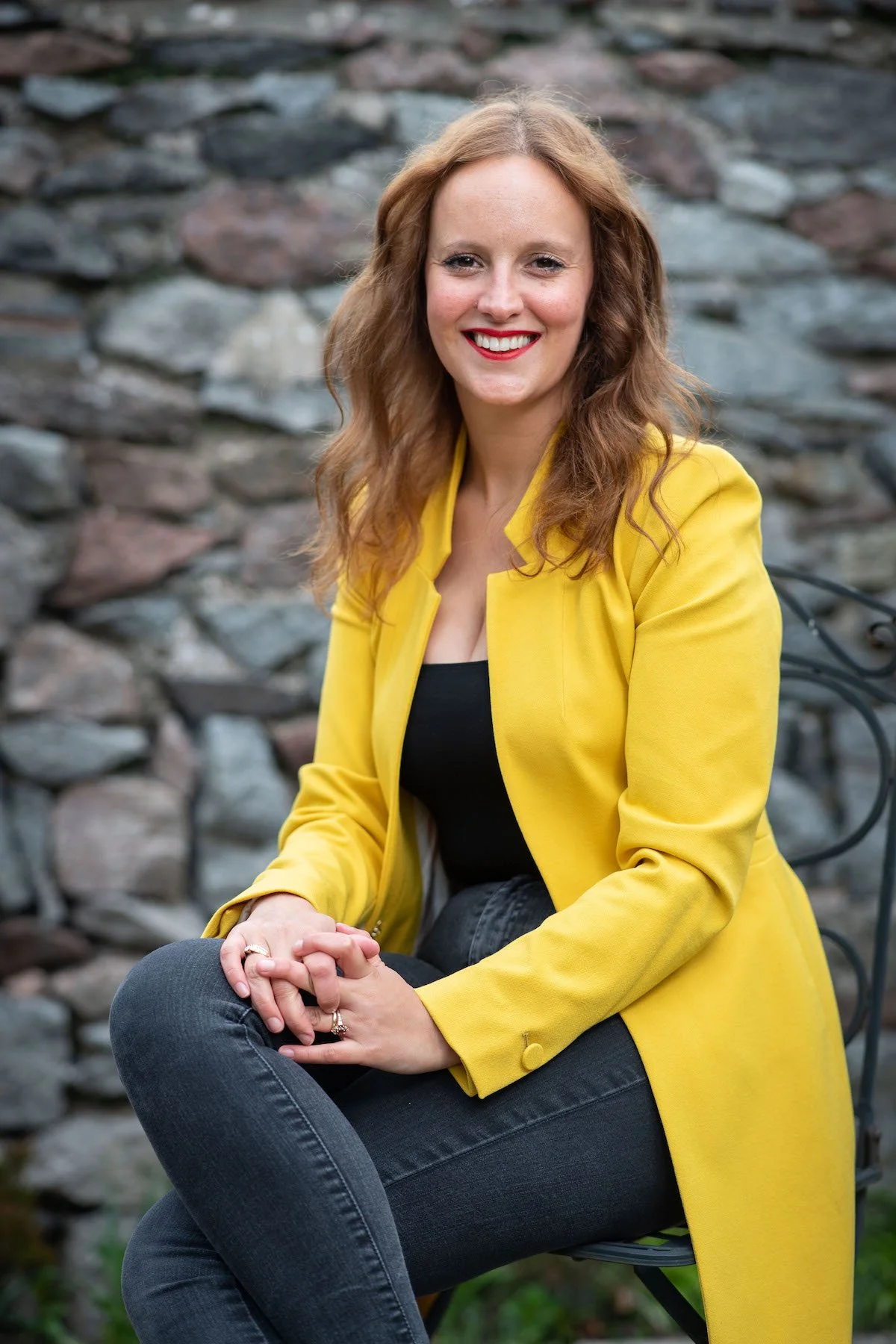 A woman with wavy red hair sitting on a metal bench outdoors, wearing a bright yellow blazer over a black top and dark jeans, smiling at the camera against a background of a stone wall.