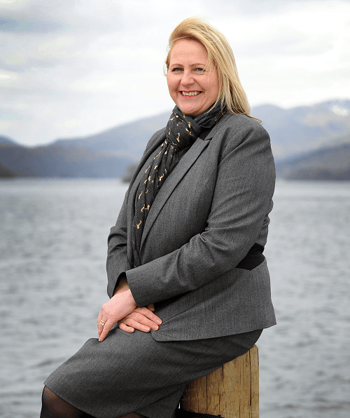 Alison Magee Barker, Founder of ImmersiV XP, in a grey business suit, sitting on a wooden post by a body of water with mountains in the background, smiling.