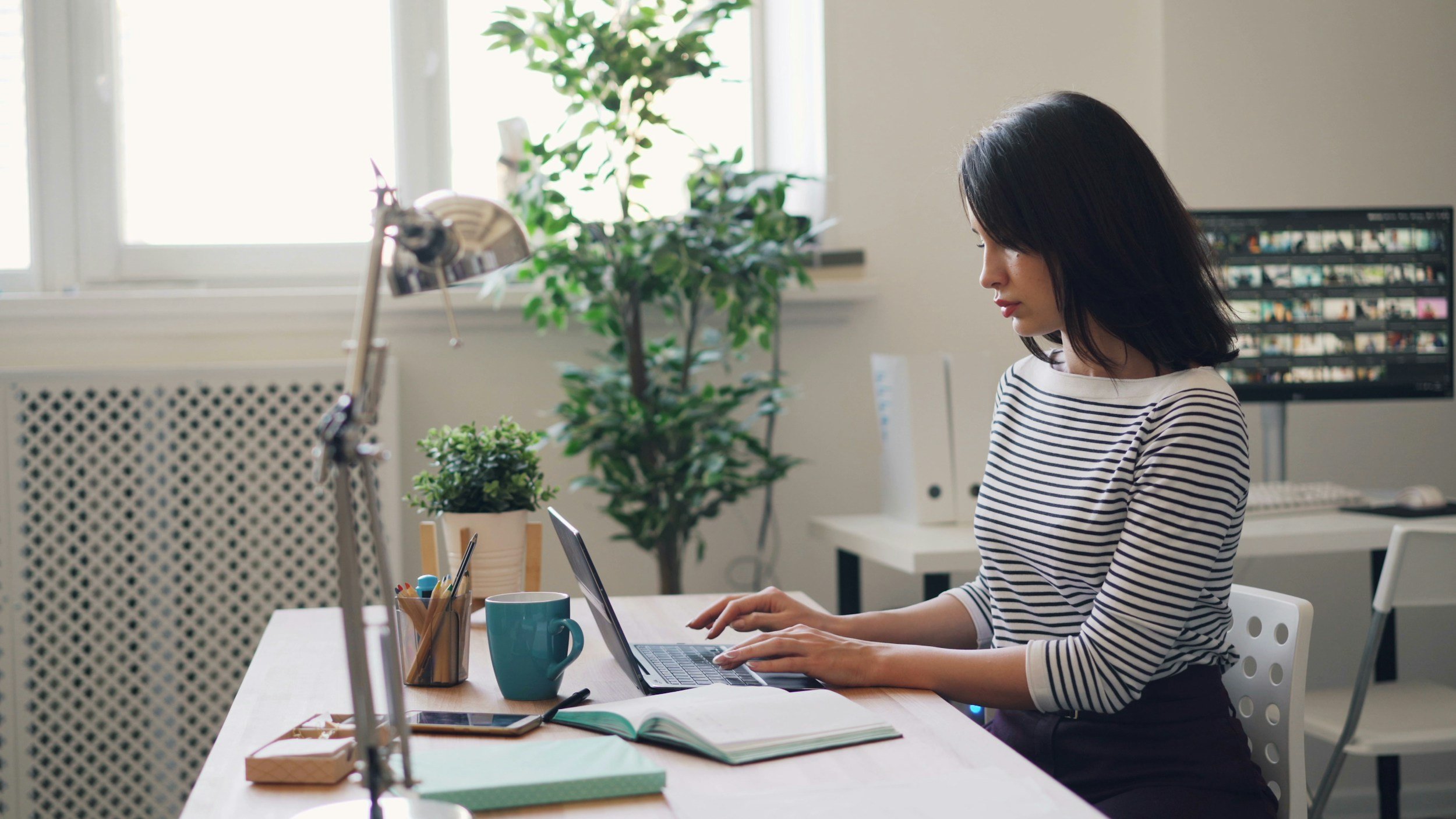 Mujer trabajando en una oficina, sentada frente a una laptop en una mesa con plantas y utensilios de oficina, con ventanas y plantas en el fondo.