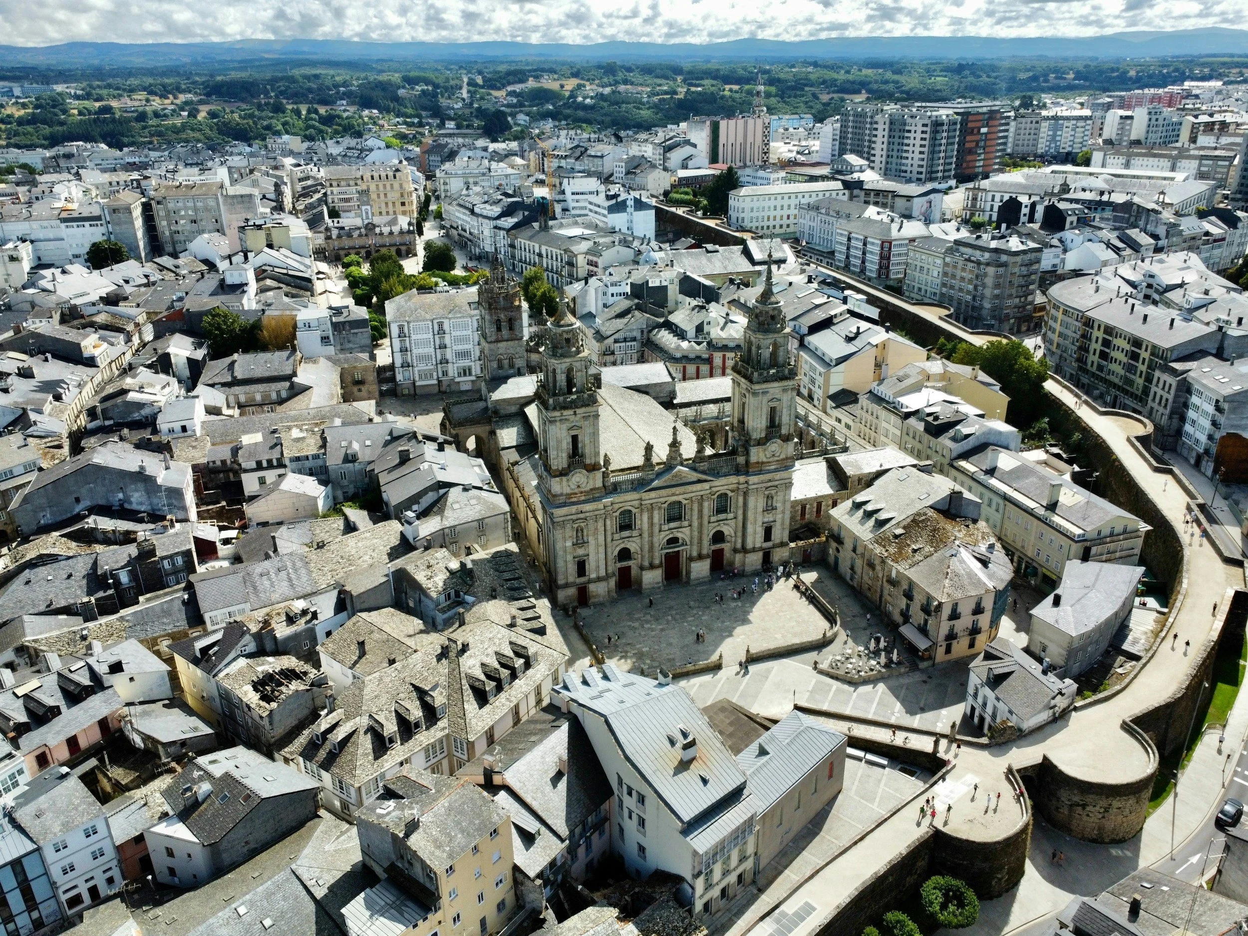 Vista aérea de una ciudad con edificios históricos y modernos, con una catedral en primer plano y montañas al fondo.
