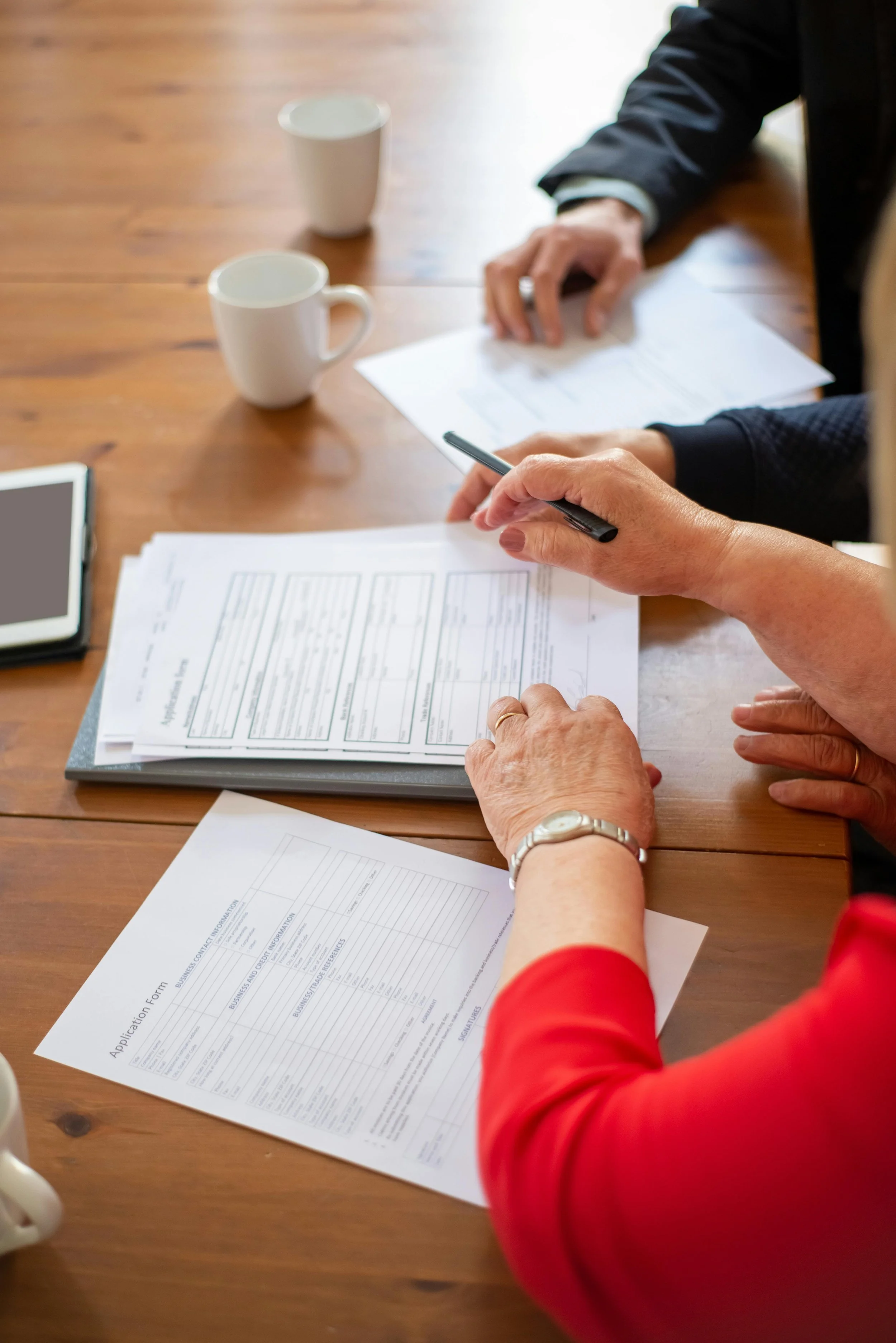 Personas revisando y llenando un formulario de solicitud en una mesa de madera con varias hojas, dos tazas blancas y un teléfono móvil visible.