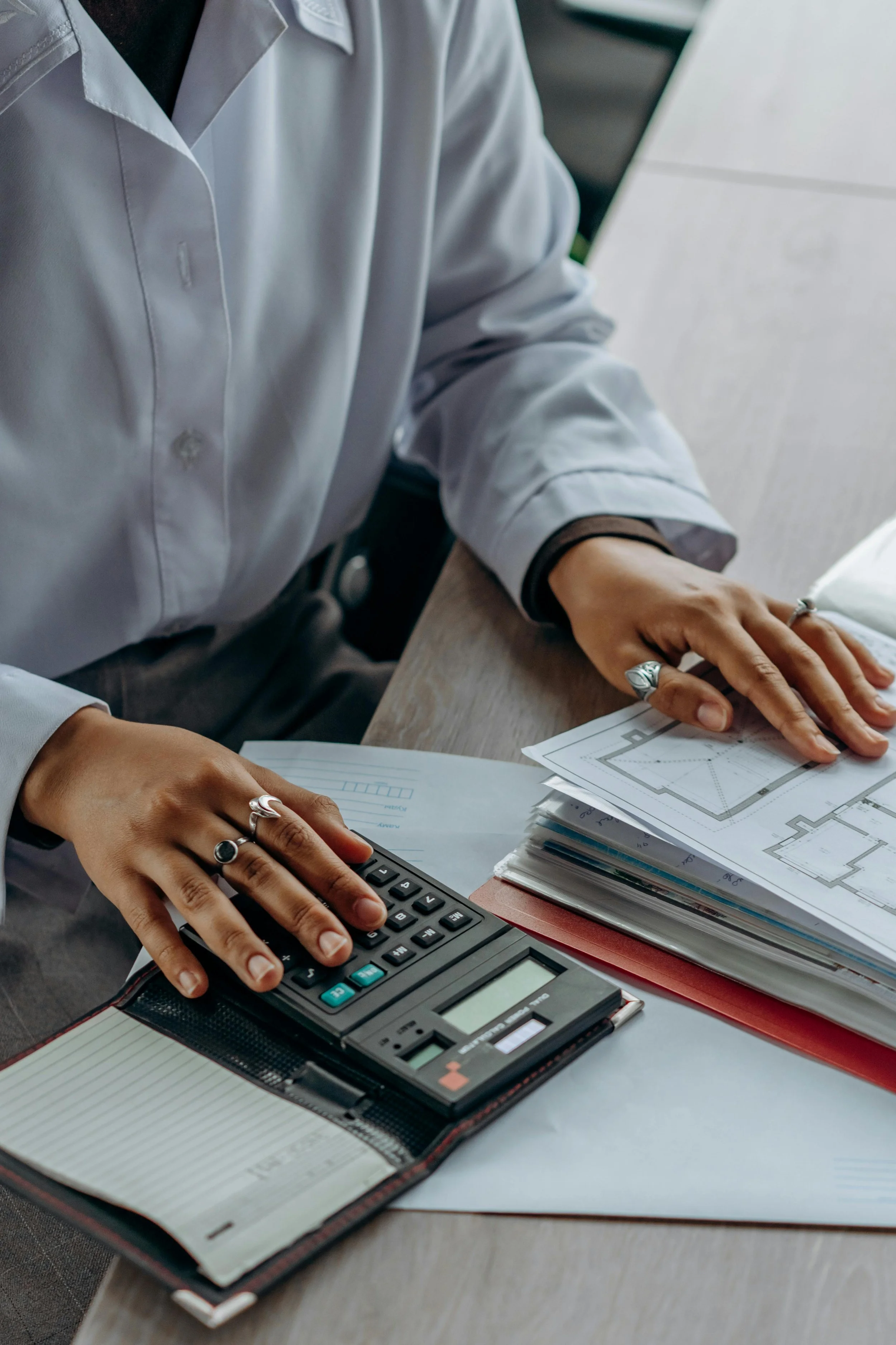 Una persona con camisón gris oscuro trabajando en una oficina, usando un apto en los dedos. Tiene un montón de papeles y documentos sobre la mesa, y está utilizando una calculadora y una computadora portátil.