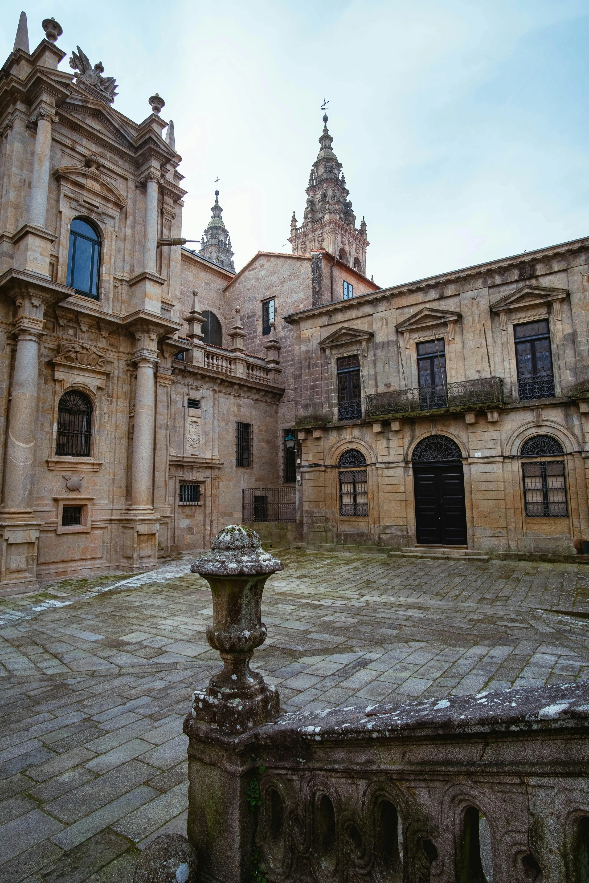 Un patio de piedra con una fuente en primer plano y edificios históricos de piedra en el fondo, con torres y ventanas ornamentadas. Cielo despejado con algunas nubes.