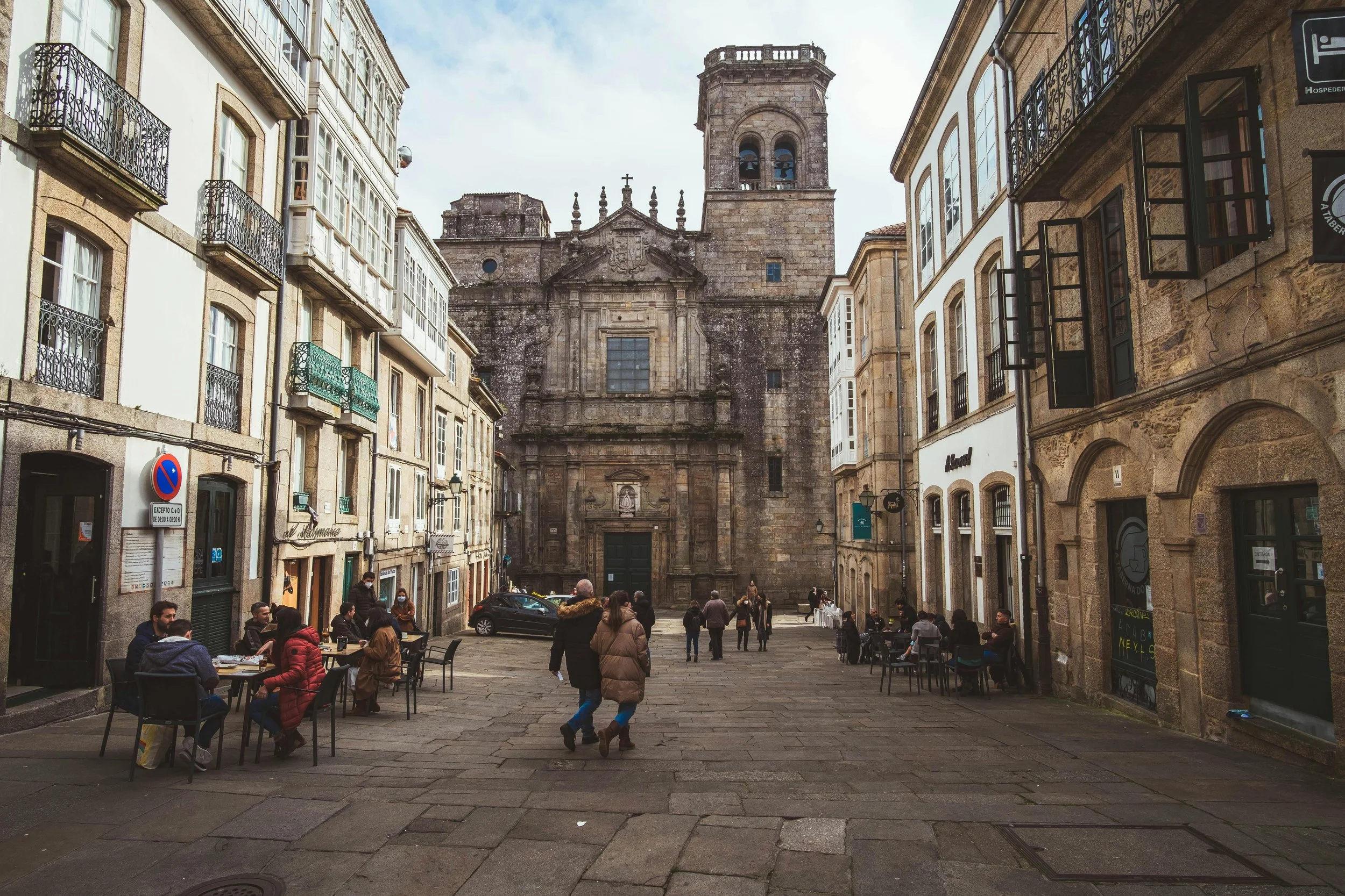 Una plaza con varias personas caminando y sentadas en mesas de restaurantes. En el fondo, una iglesia antigua de piedra con campanario y detalles arquitectónicos tradicionales. Edificios de varias plantas rodean la plaza.