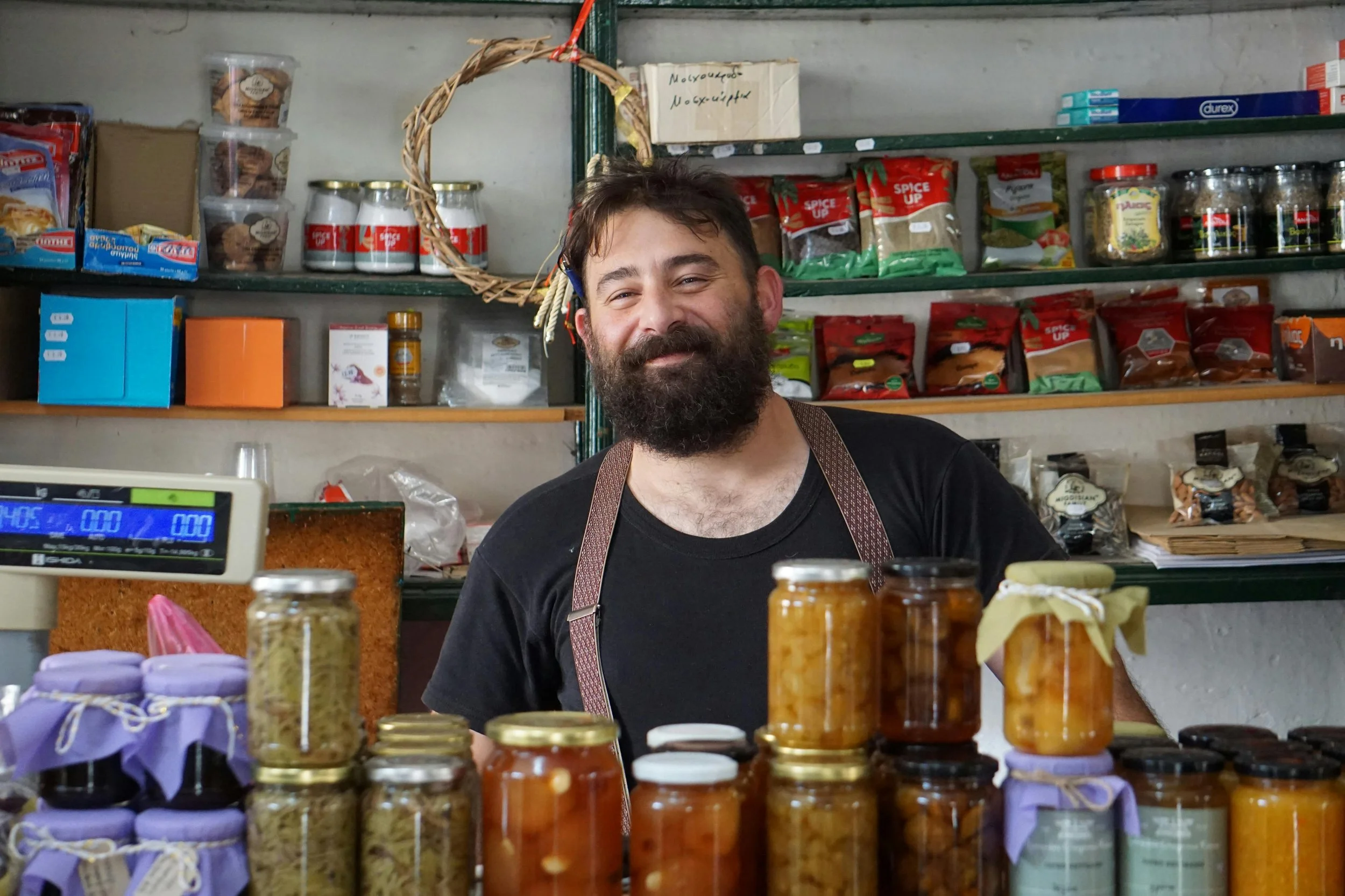 Hombre con barba sonriendo en una tienda de abarrotes, rodeado de frascos de mermeladas y productos en estantes.