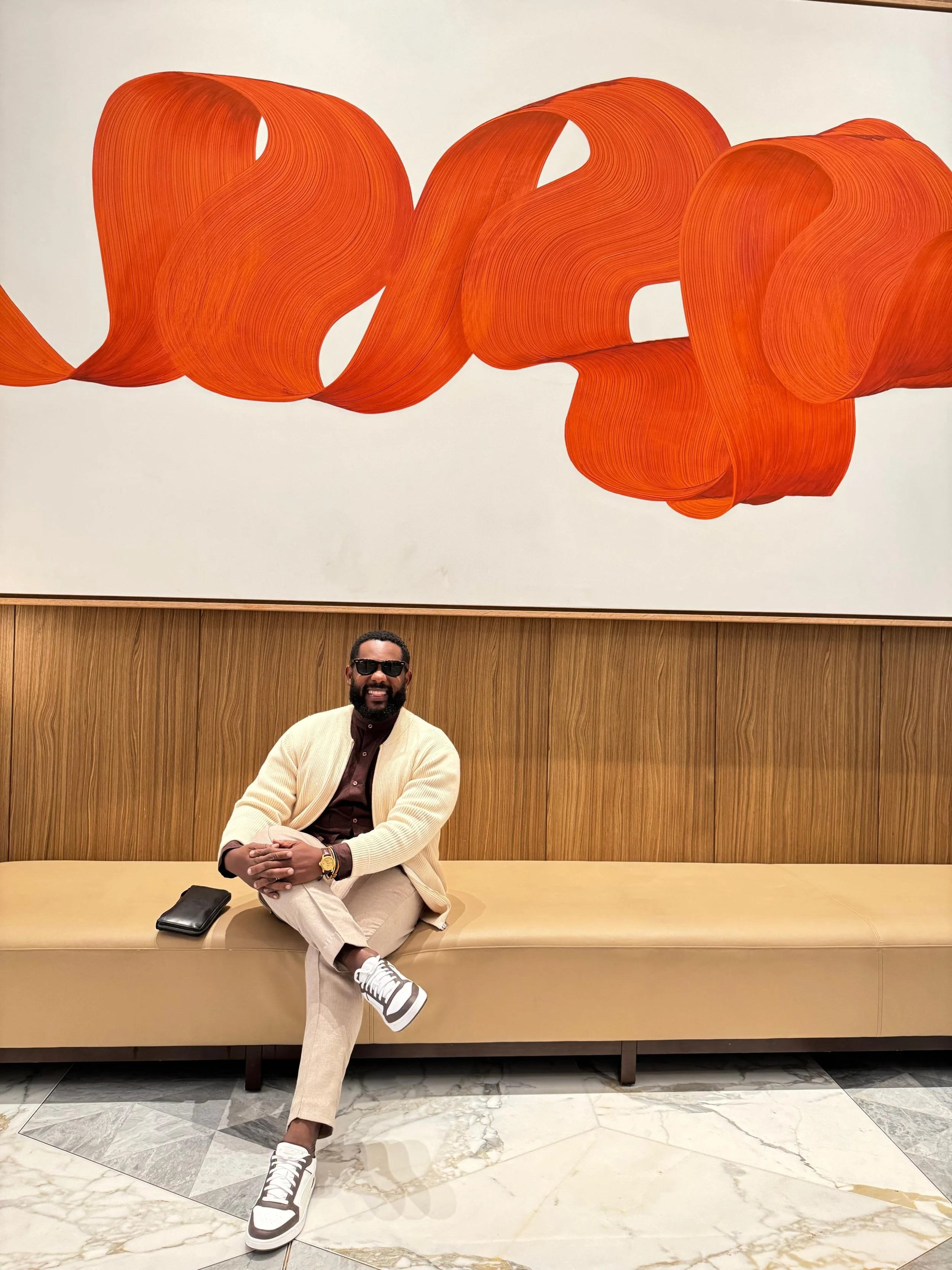 A man with dark skin, wearing sunglasses, a beige cardigan, a dark shirt, light-colored pants, and sneakers, sitting on a beige bench in front of a large colorful abstract art piece with orange swirls. The setting appears to be a modern indoor space with marble flooring.
