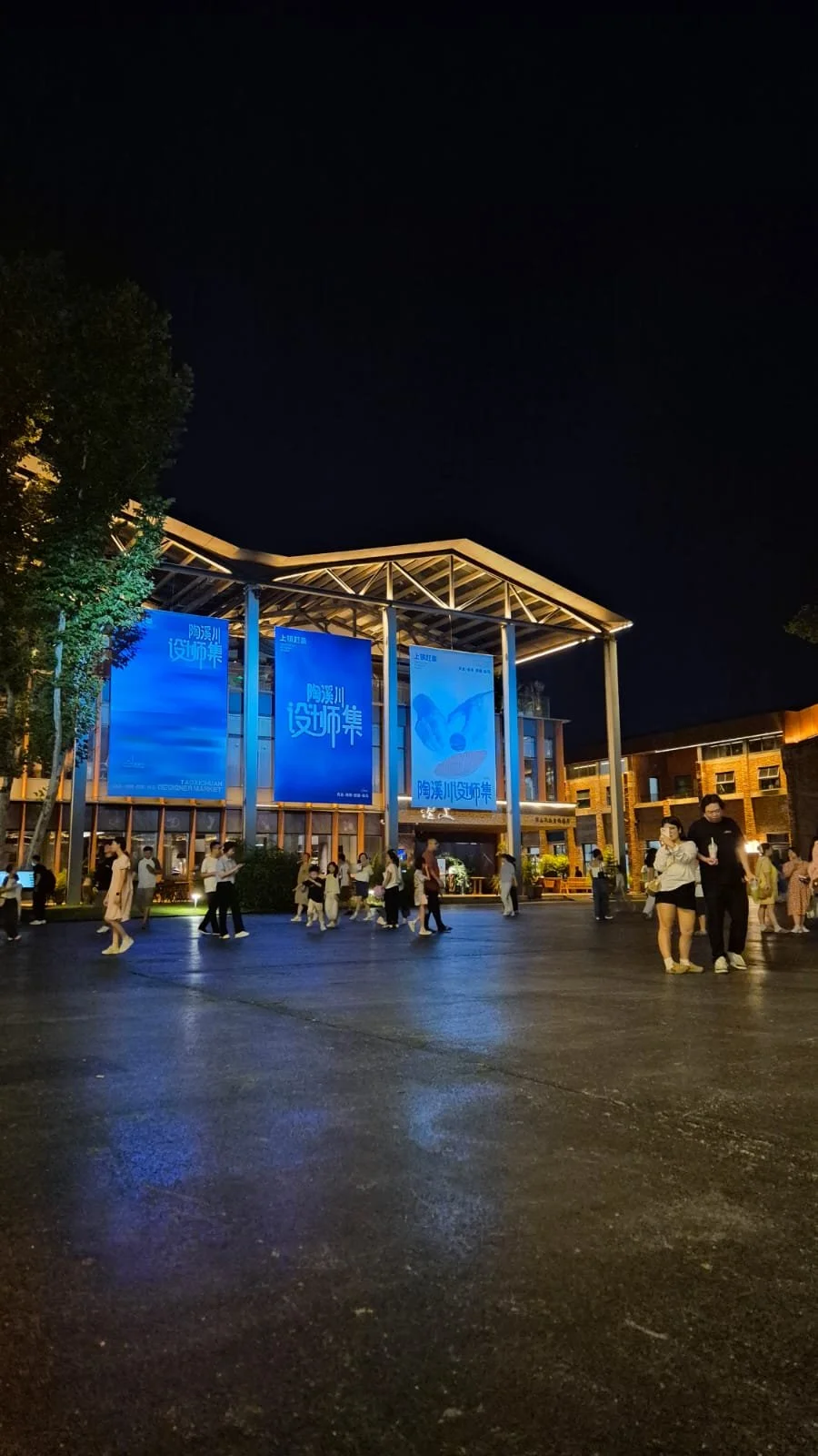 Night scene at a public square with people walking and gathering under a modern open structure with large blue banners displaying Chinese characters, surrounded by trees and buildings.