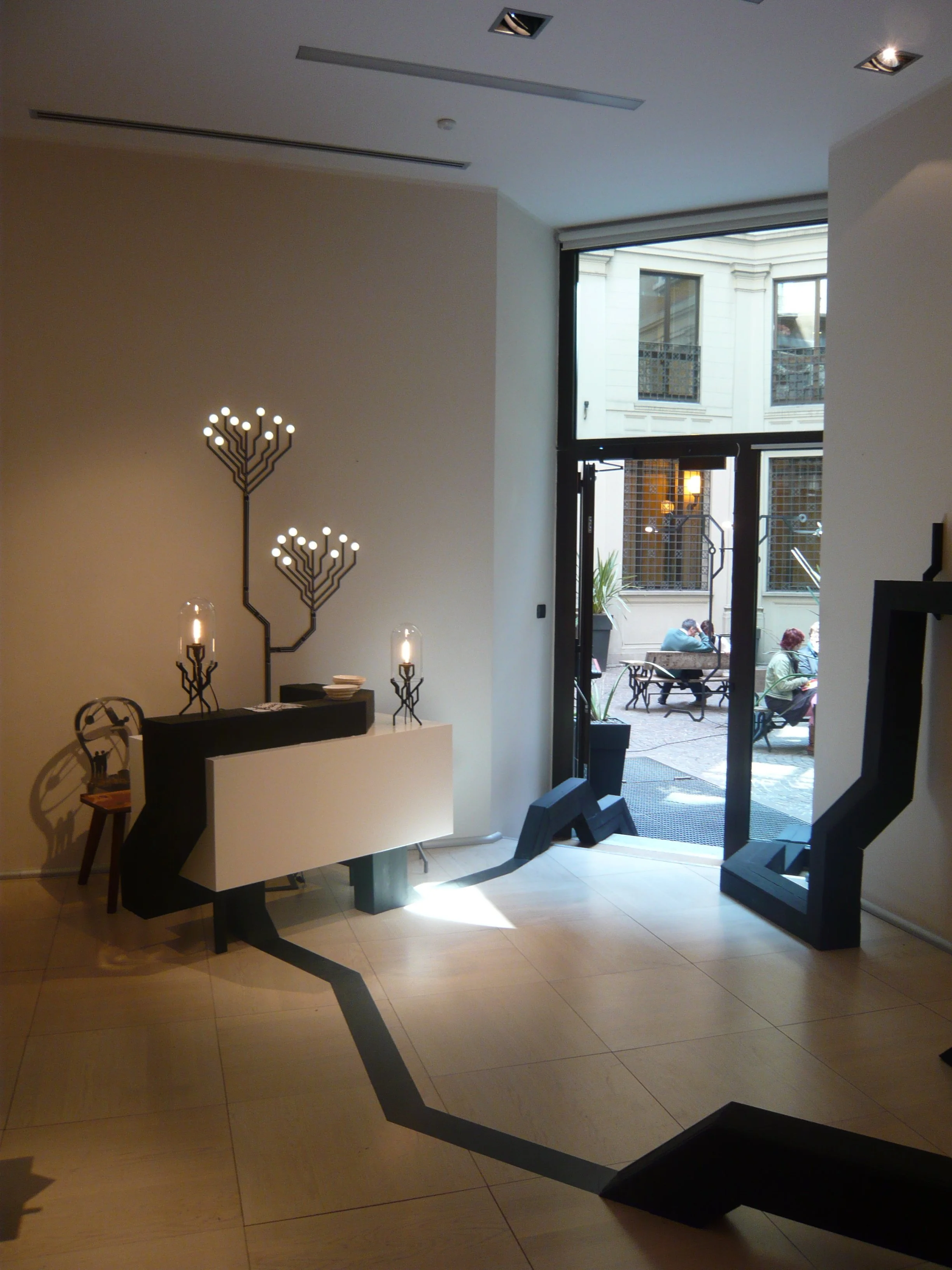 Interior view of a modern hotel lobby with black and white decor, featuring artistic black furniture with geometric shapes and a distinctive black branch-like lamp sculpture. Sunlight enters through a large glass door leading to an outdoor seating ar