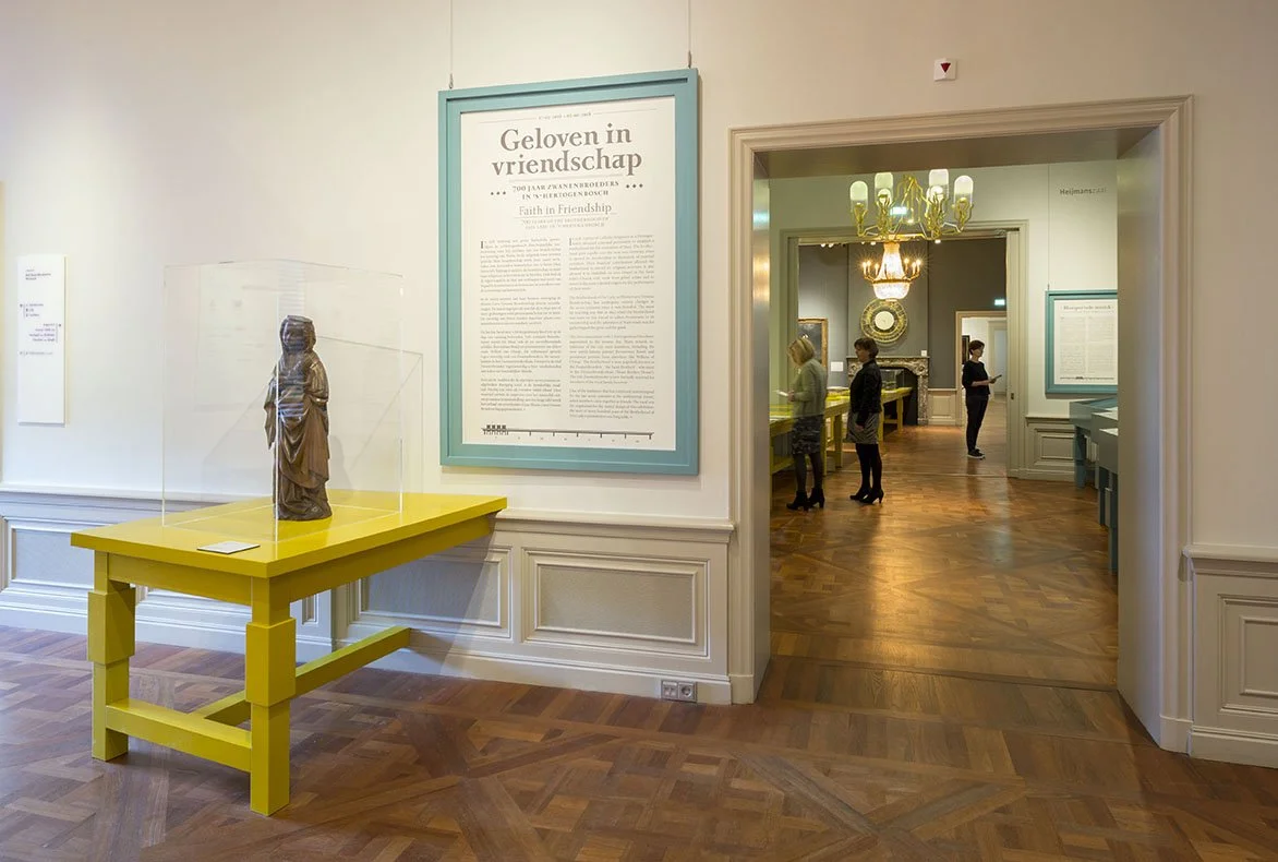 An art gallery interior showing a small sculpture on a bright yellow table inside a glass case, with framed information outside the case and a large framed informational sign on the wall. In the background, there are visitors looking at artworks, cha