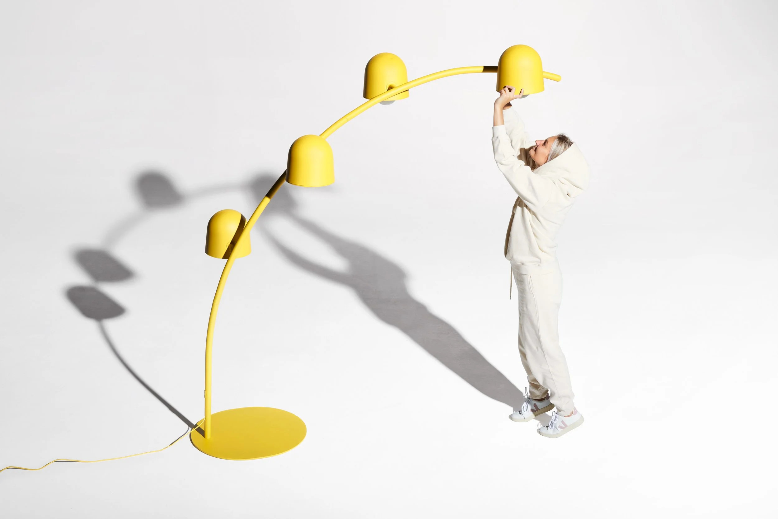 A woman in a white hoodie and pants adjusting a yellow modern arc floor lamp against a plain white background.