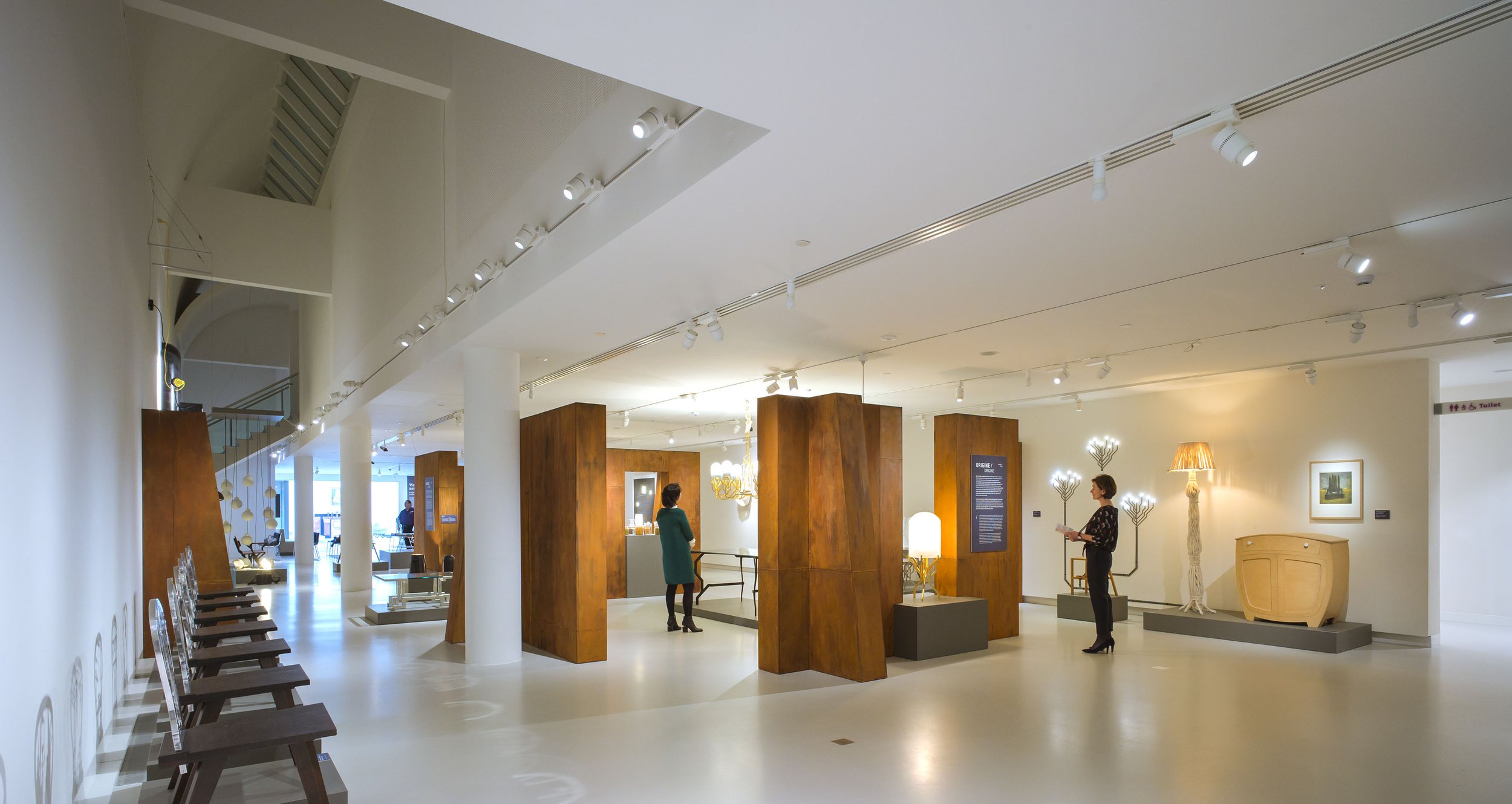 Art gallery interior with wooden display panels, sculptures, lamps, and visitors viewing artwork.