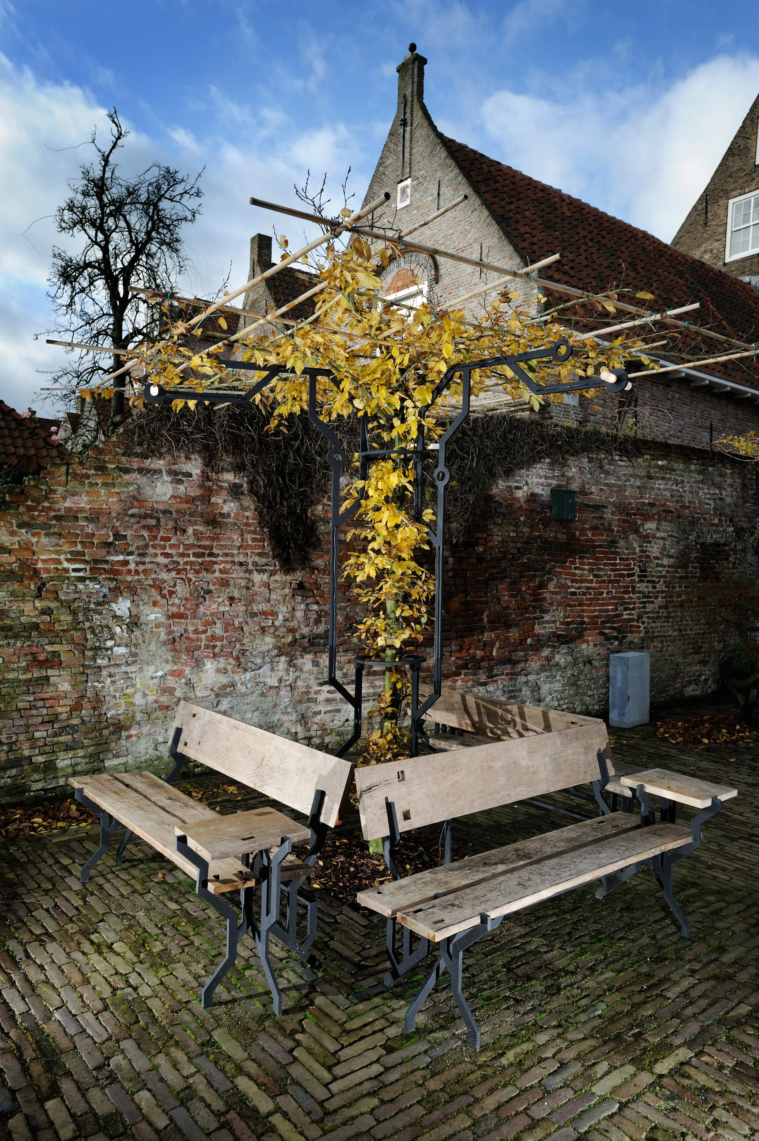 Outdoor scene with a metal tree sculpture wrapped with yellow leaves, surrounded by four wooden benches on a cobblestone pathway, brick wall, and old buildings in the background.