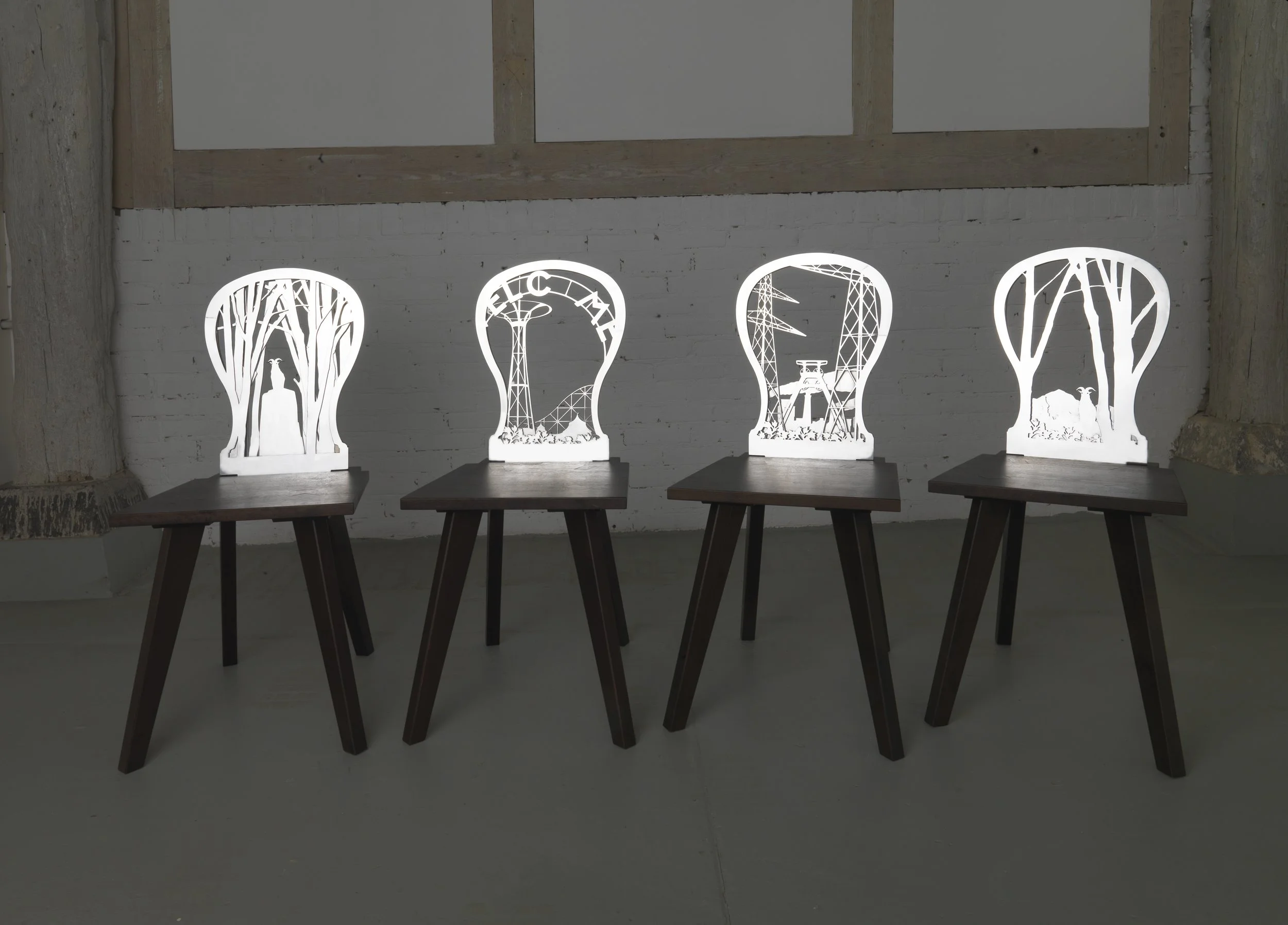 Four wooden chairs with white backrests featuring industrial and nature-inspired cutout designs, arranged on a light gray floor against a white brick wall.