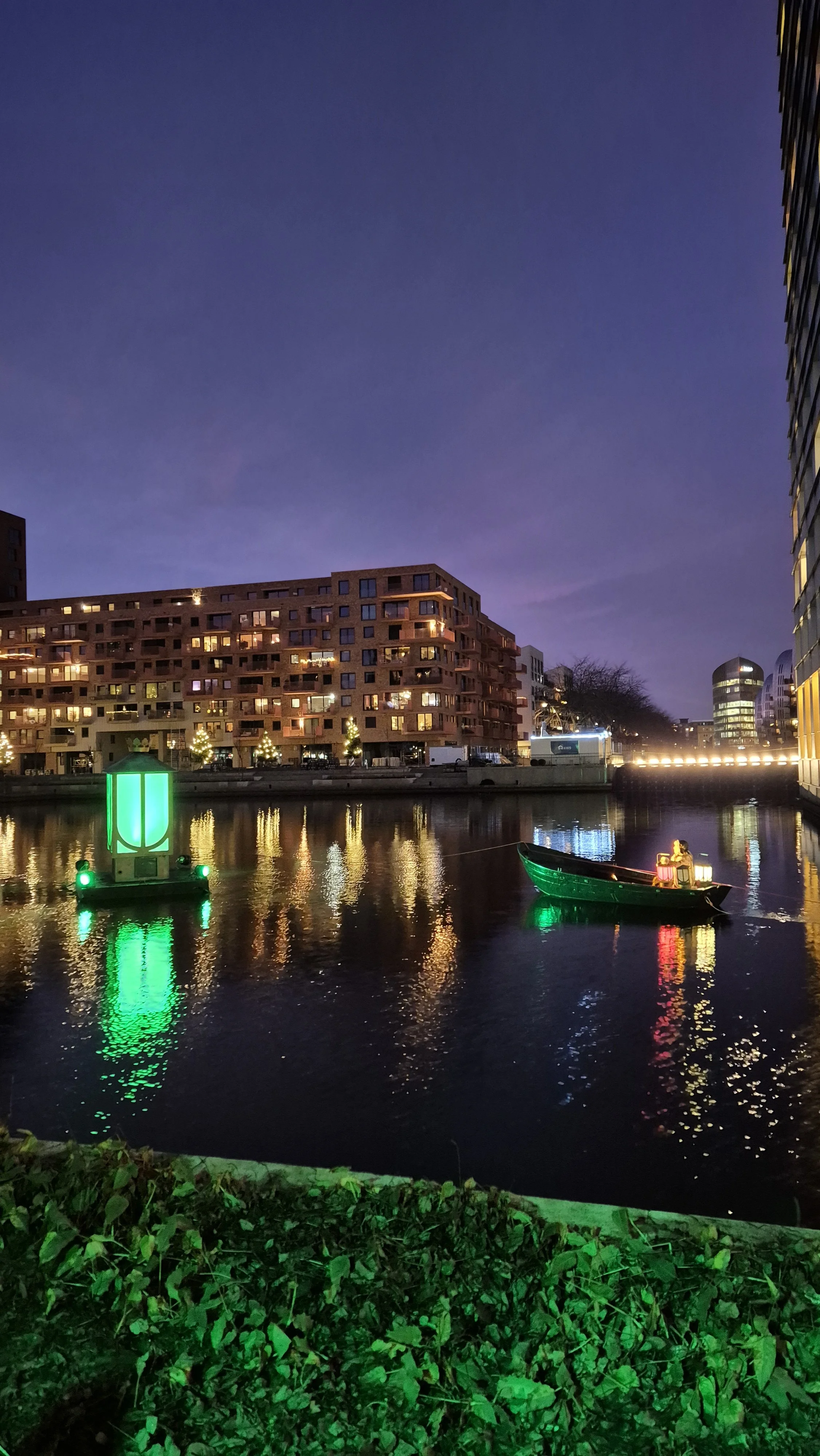A nighttime scene of a lit cityscape reflections on a river, with a small boat carrying two passengers, and green and yellow lights on buildings and water.