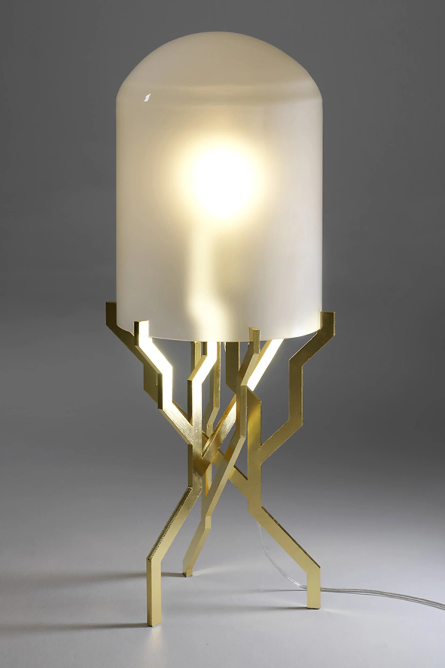 A modern table lamp with a cylindrical, frosted white glass shade and a gold geometric metal base, turned on and emitting a warm glow.