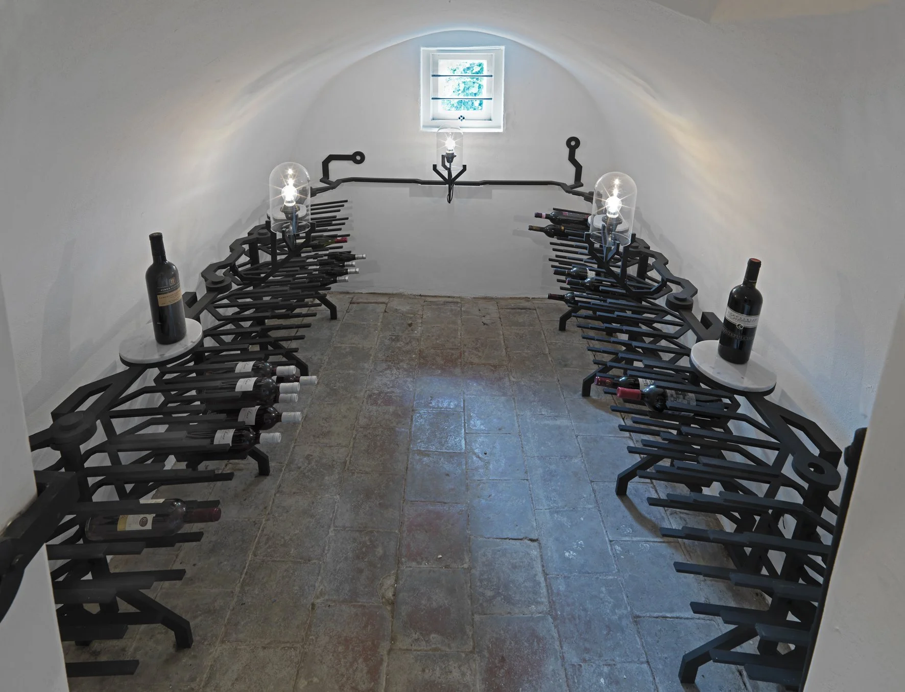 A wine cellar with curved walls and stone flooring. Two black metal wine racking units form a U-shape, holding various bottles of wine and a few glasses. On each end of the wine racks, there are small round white tables with bottles of wine on them. 