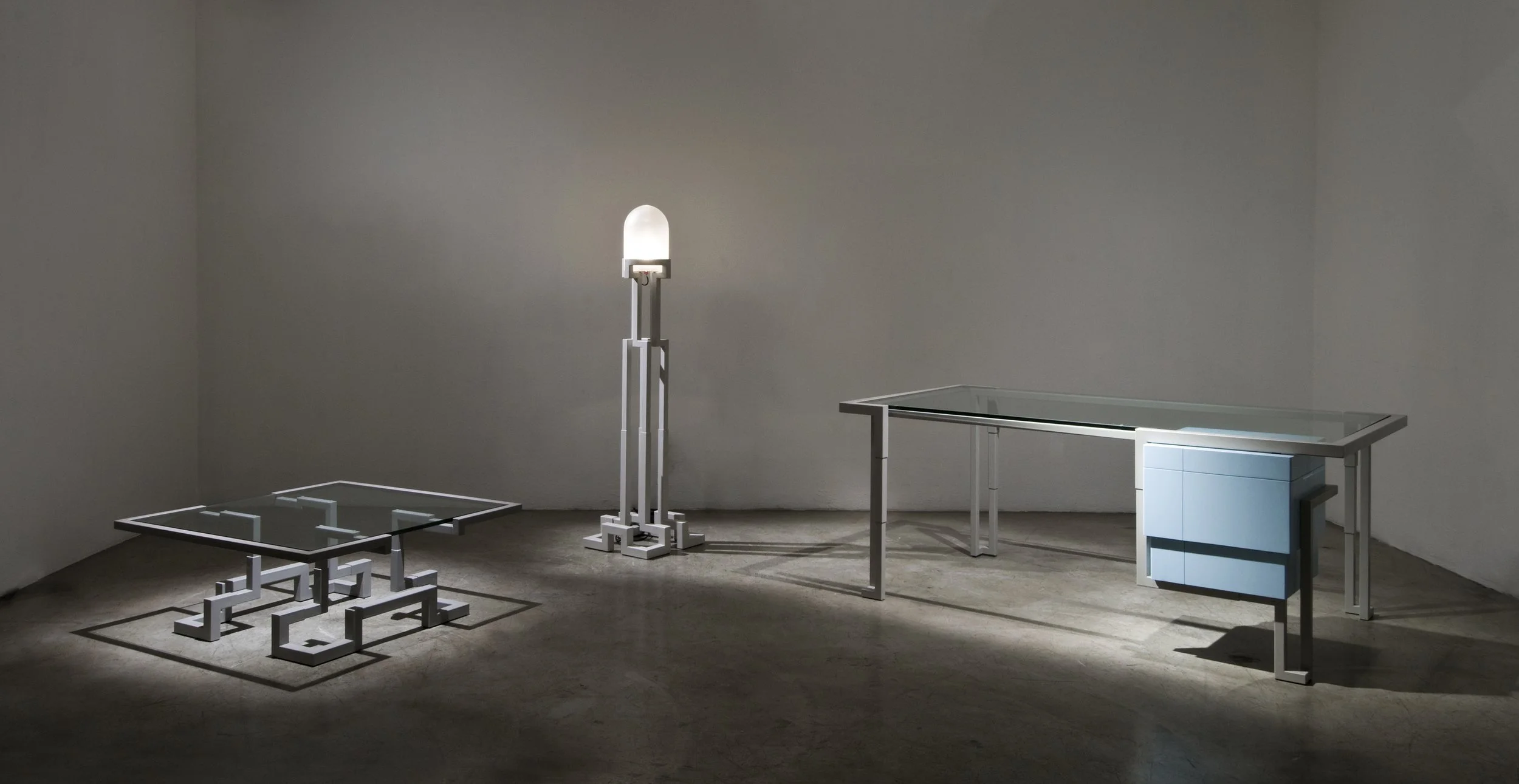 Minimalist modern furniture including a glass-top coffee table, a desk with a light blue drawer unit, and a tall floor lamp, all with metallic frames, arranged in a sparsely decorated room with concrete floors and plain white walls.