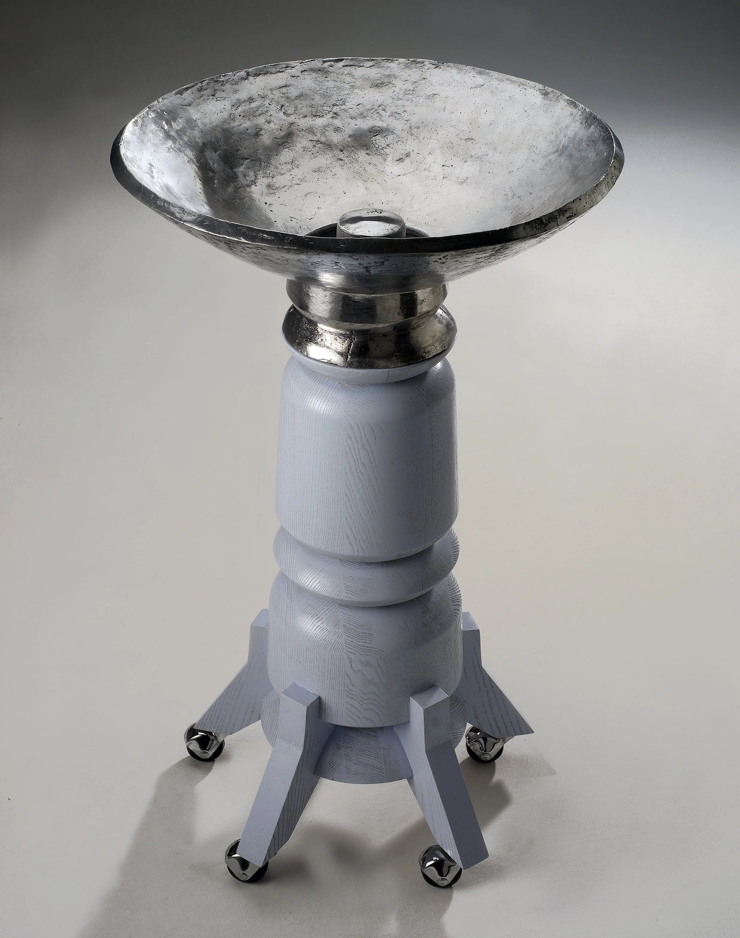A modern, artistic table with a silver bowl on top, a white wooden base with wheels, and a minimalist design.