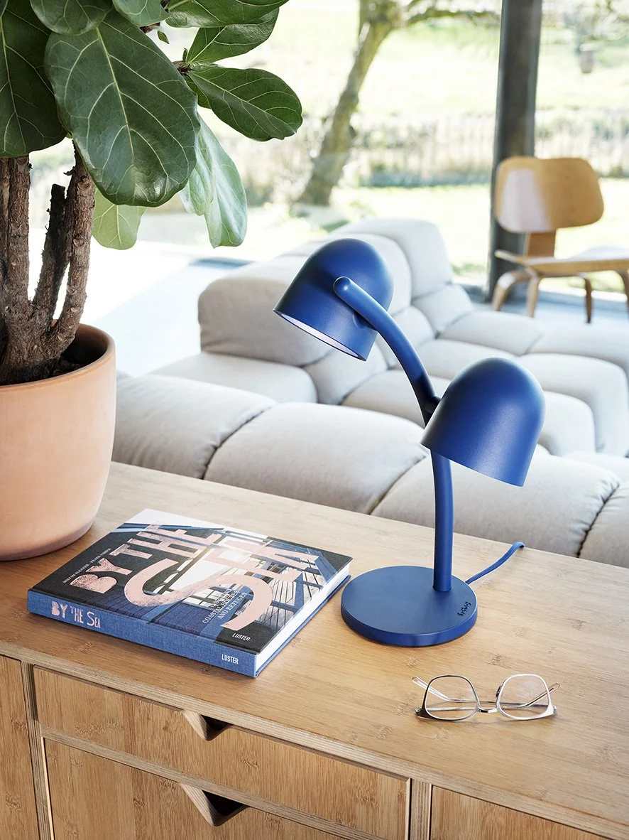 Blue double-headed desk lamp on a wooden side table with a book titled 'BY THE SEA,' a pair of glasses, and a large houseplant in a terracotta pot. In the background, a beige sofa and a wooden chair are visible in a sunlit living room.