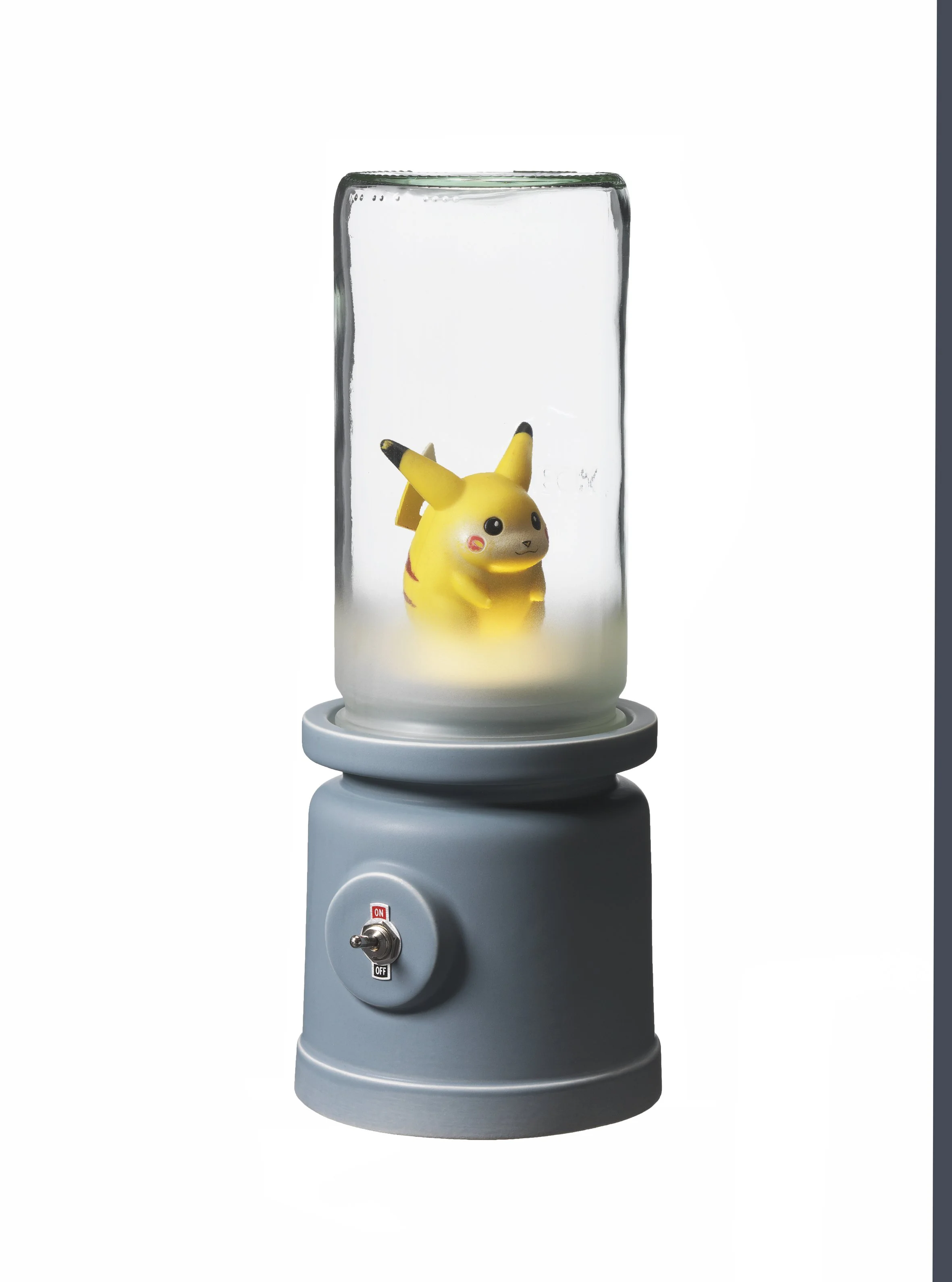 A small Pikachu figurine inside a transparent cylindrical display case on a gray stand with a switch at the front.