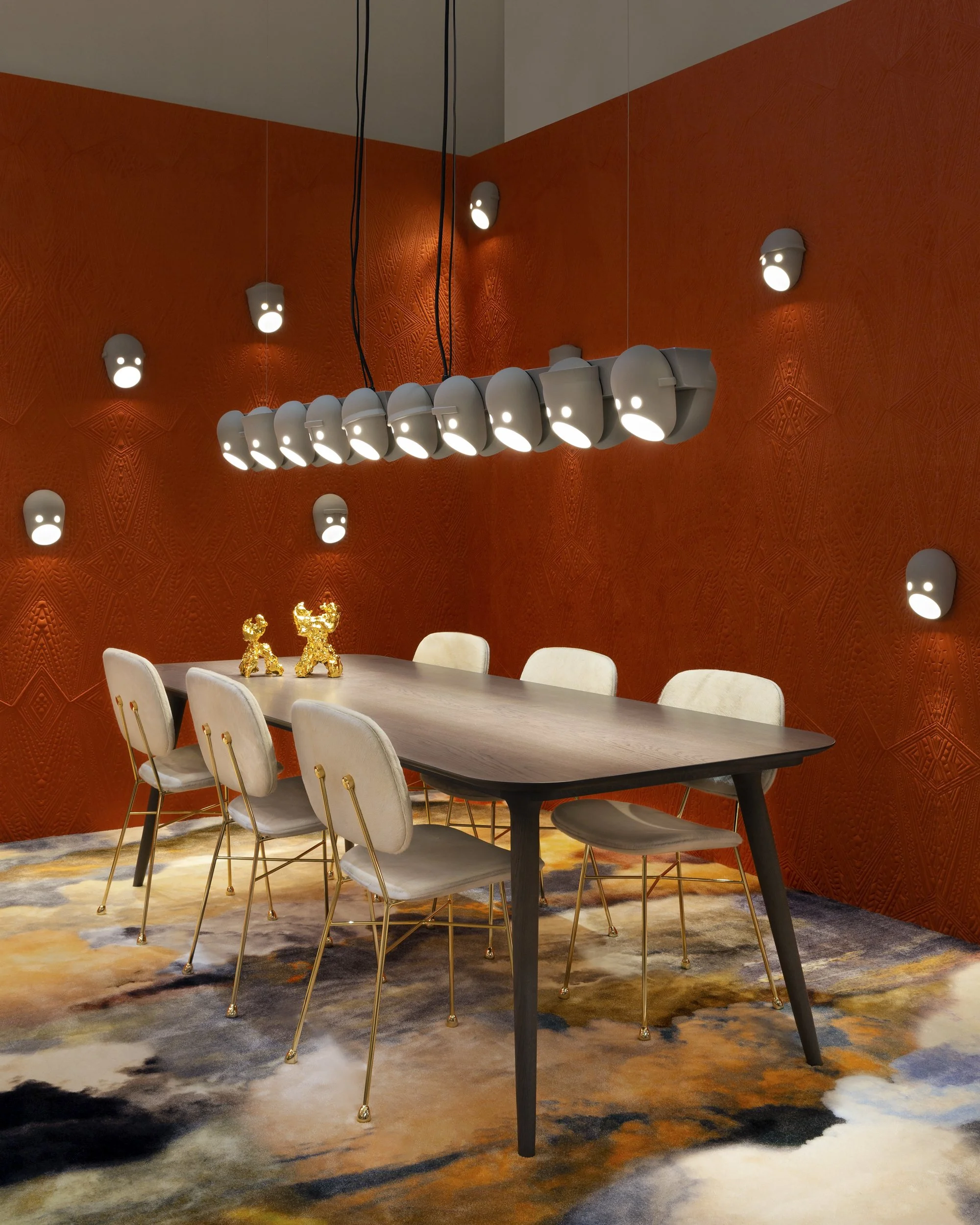 Modern dining room with a wooden table, white cushioned chairs with gold legs, orange textured walls with wall-mounted lights, and a contemporary chandelier with multiple rounded lights. Gold reindeer decorations are on the table, and a colorful abst