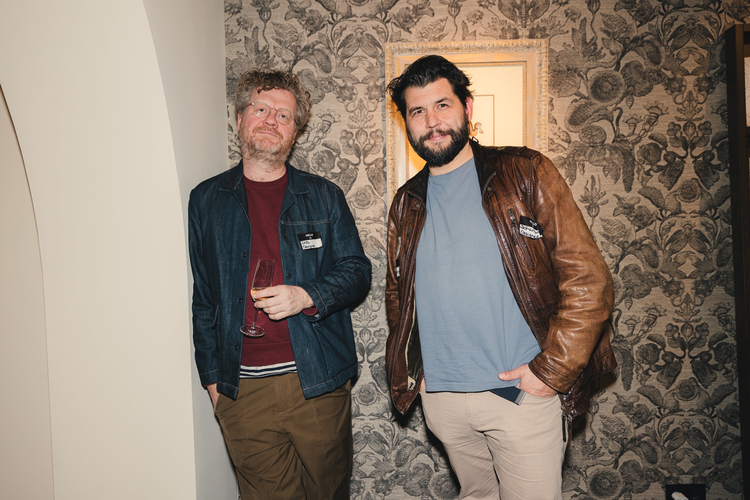 Two men standing together at a social event, one holding a glass of champagne, in front of a floral-patterned wallpaper and a framed mirror.