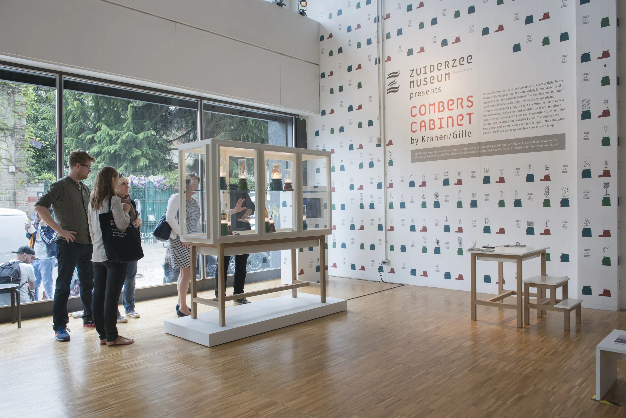 Interior of an art museum gallery with a group of people viewing an exhibit of a glass display case on a wooden table. The display case contains ceramic objects shaped like containers. Large windows with a view of trees and people outside are visible