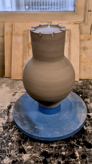A ceramic vase on a pottery wheel in a workshop, with a wooden window and tools in the background.
