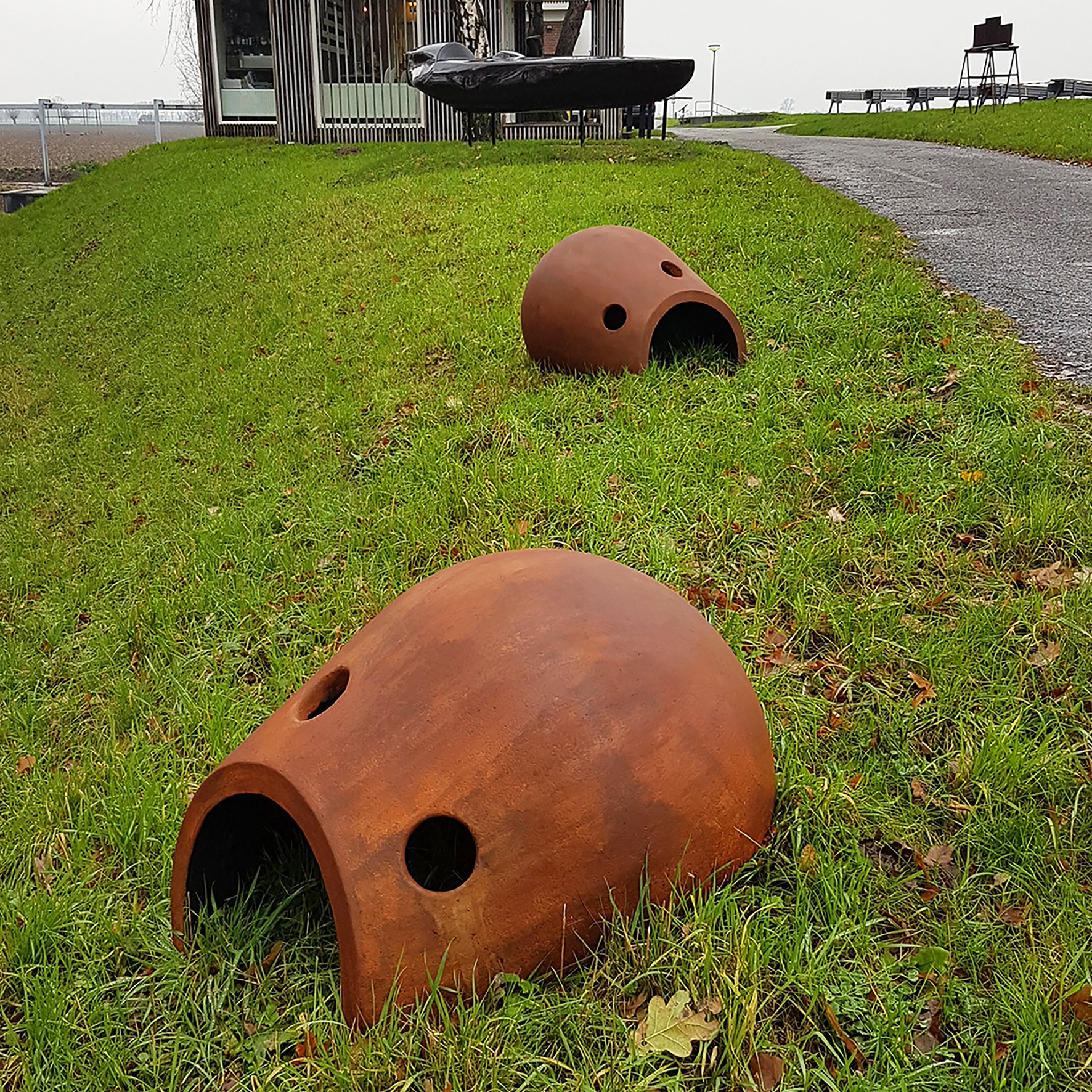 Two large, rust-colored, dome-shaped sculptures with circular openings, lying on a grassy area beside a paved pathway. One sculpture is in the foreground, and the other is further back, near a bench and a small building.