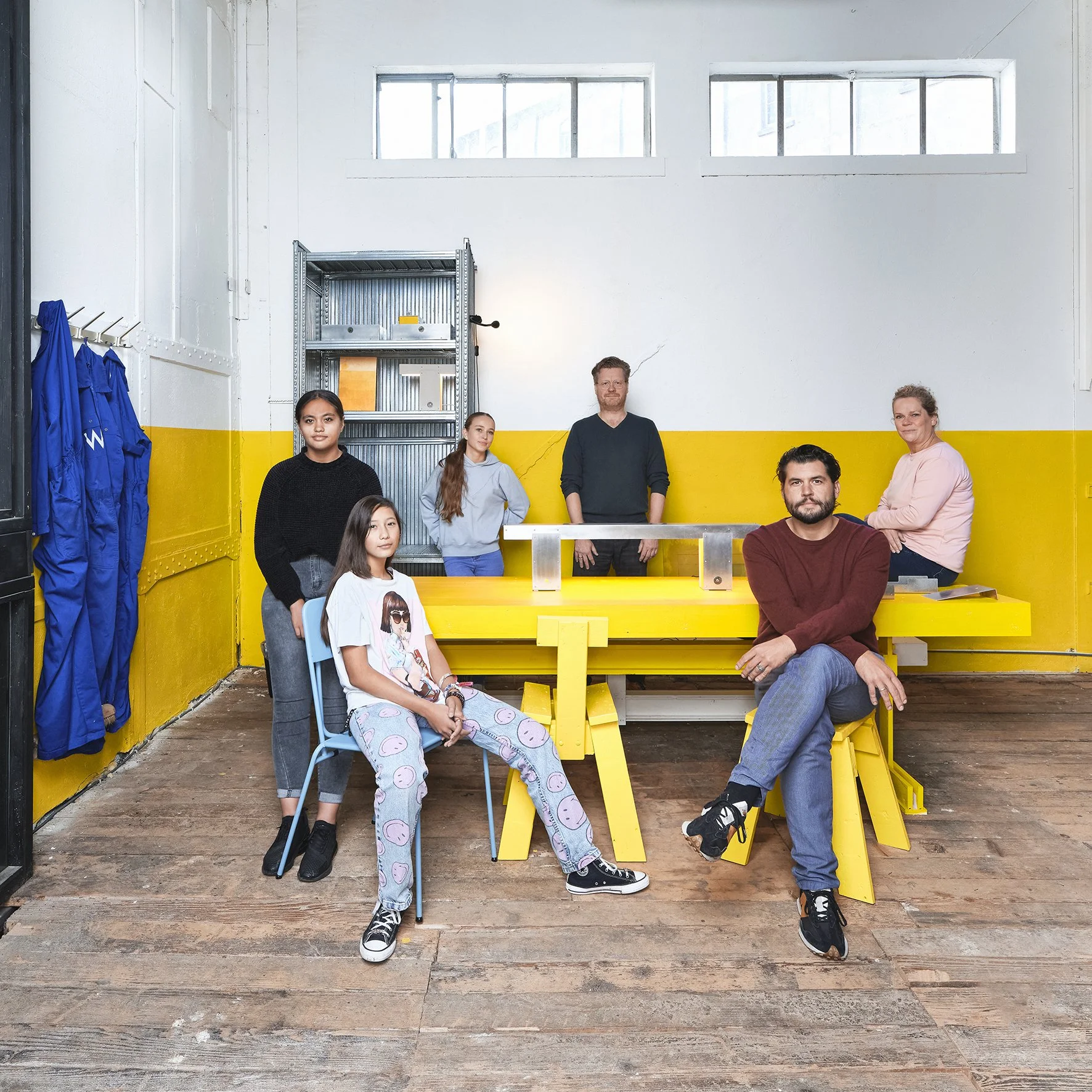 Group of six people in a room with a yellow and white wall, some sitting and some standing, with a yellow table and metal shelving in the background.
