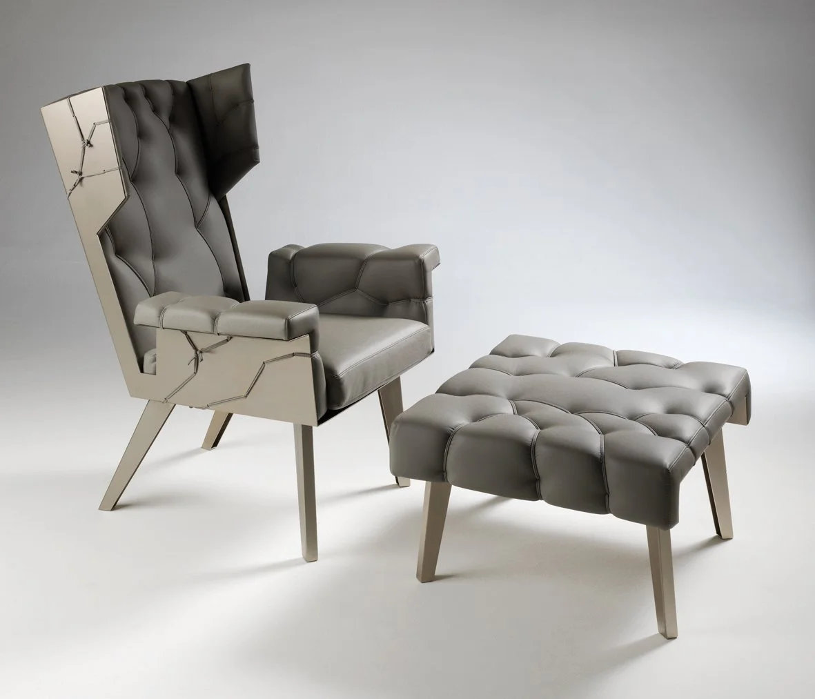 A modern leather armchair with a matching tufted ottoman, both with light wooden legs, against a plain gray background.