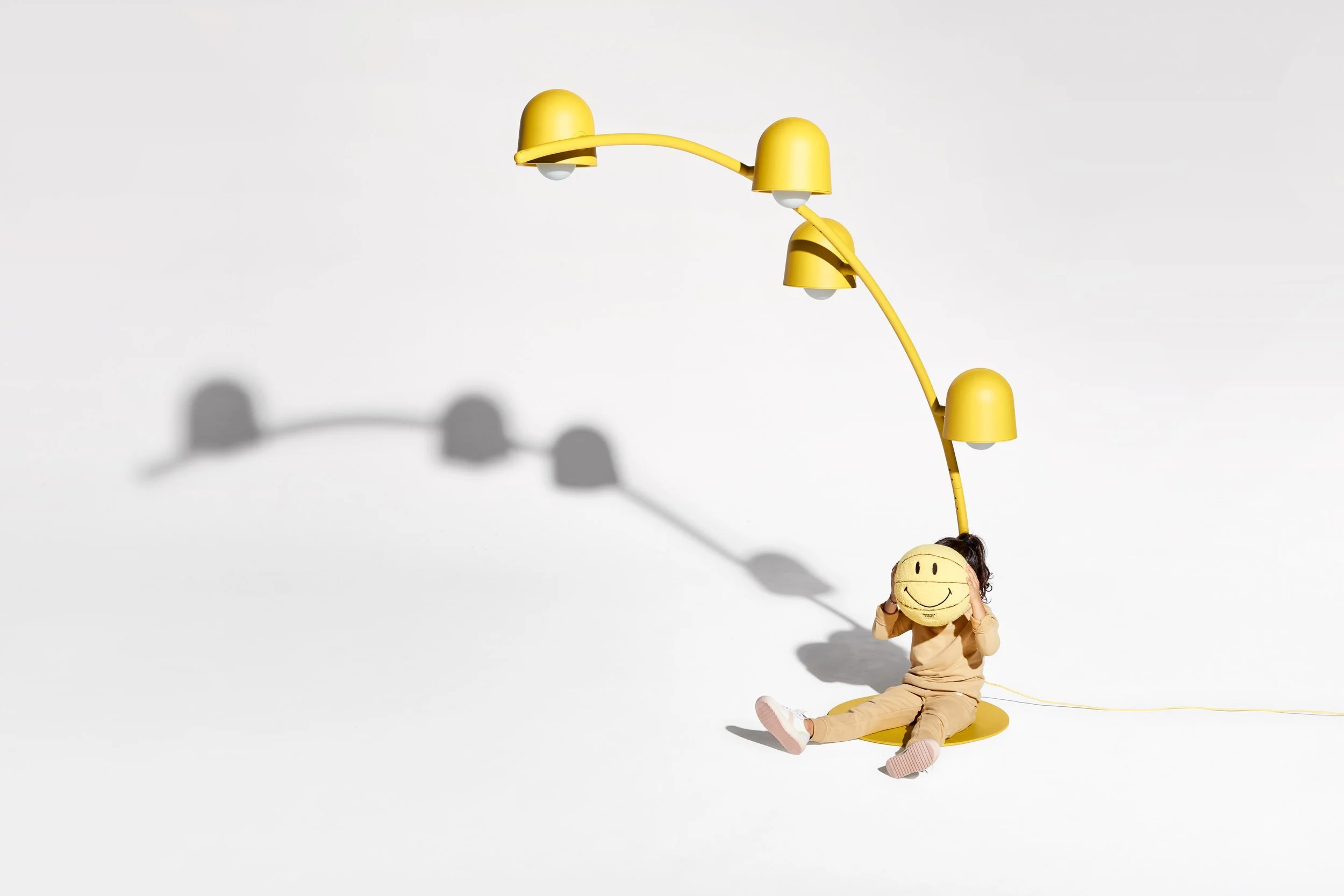 Child sitting on the floor holding a smiley face pillow, with a yellow modern desk lamp overhead casting a shadow, against a plain white background.