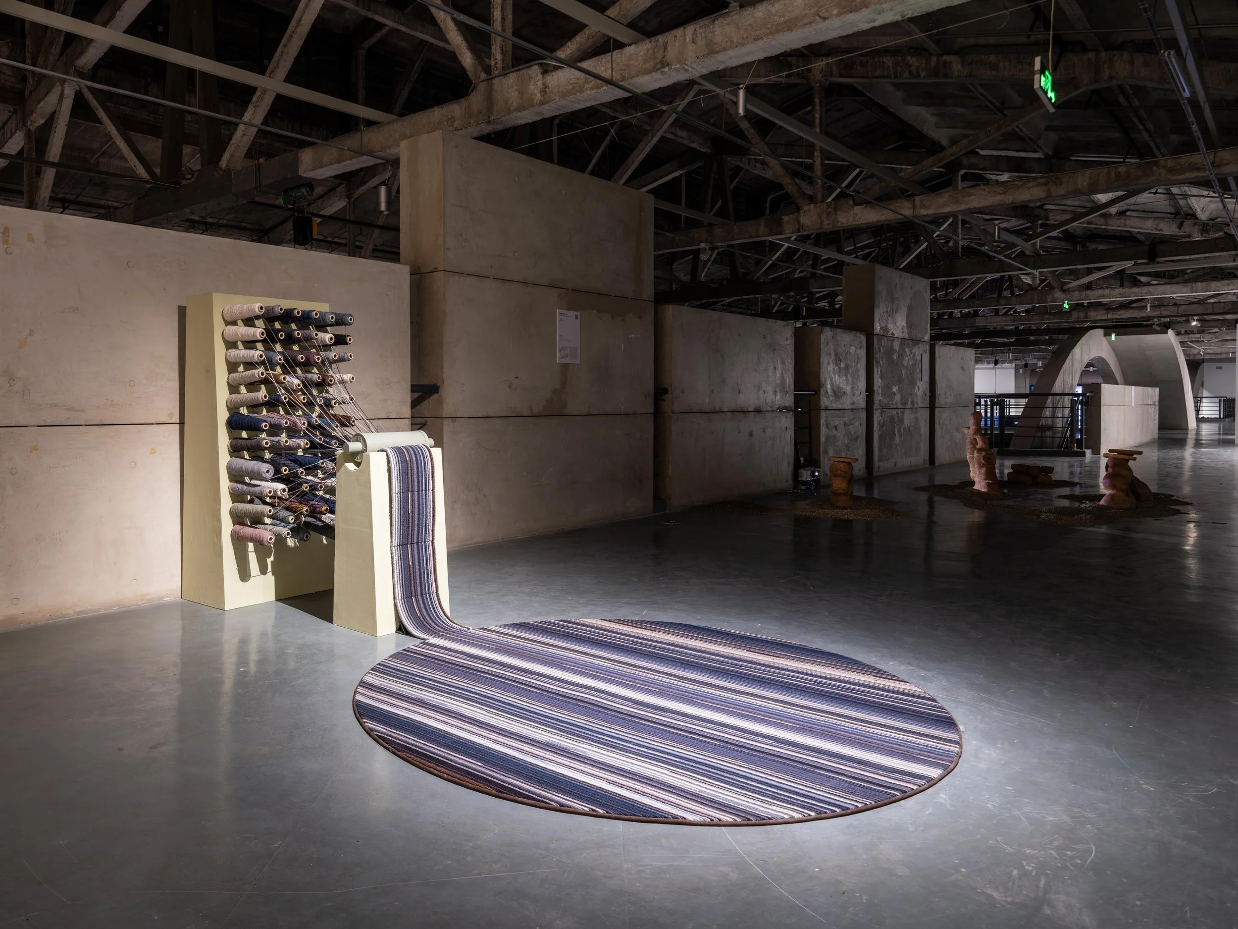 An art installation featuring a vertical structure made of spools of thread with some threads extending onto a striped circular rug on the floor. The setting appears to be a spacious, industrial-style gallery with concrete walls and exposed ceiling b