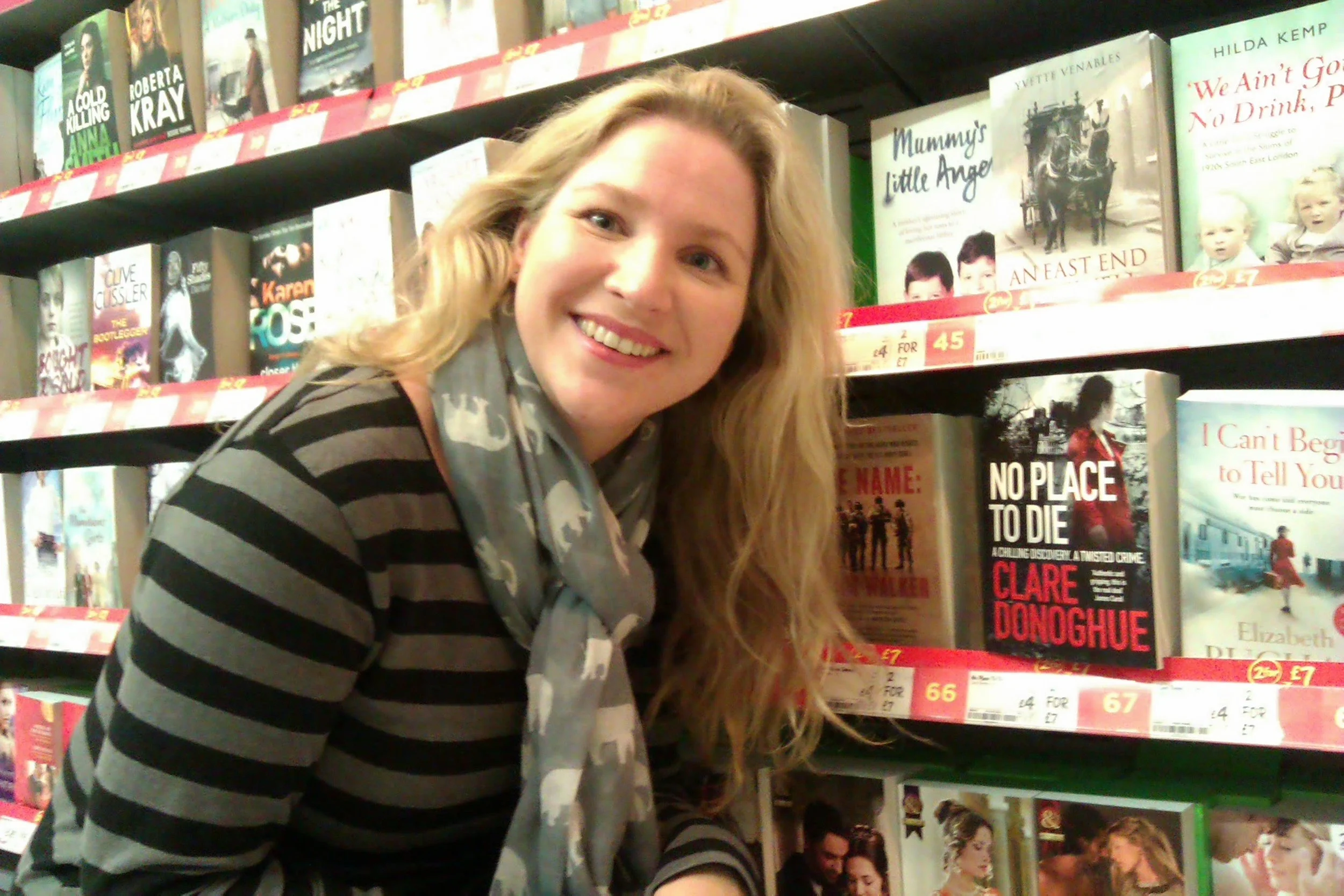 A woman with long blonde hair and a striped shirt smiling in a bookstore aisle with shelves of books behind her.