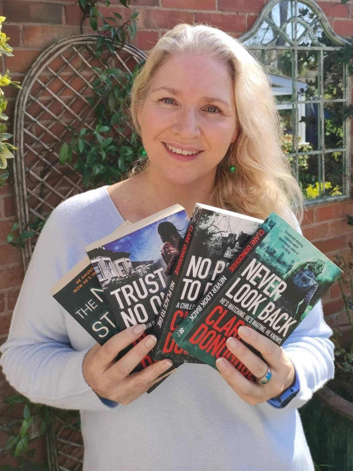 A smiling woman with blonde hair holding several books titled 'Trust No One,' 'No Place to Sleep,' and 'Never Look Back,' standing outdoors in front of a brick wall and garden.