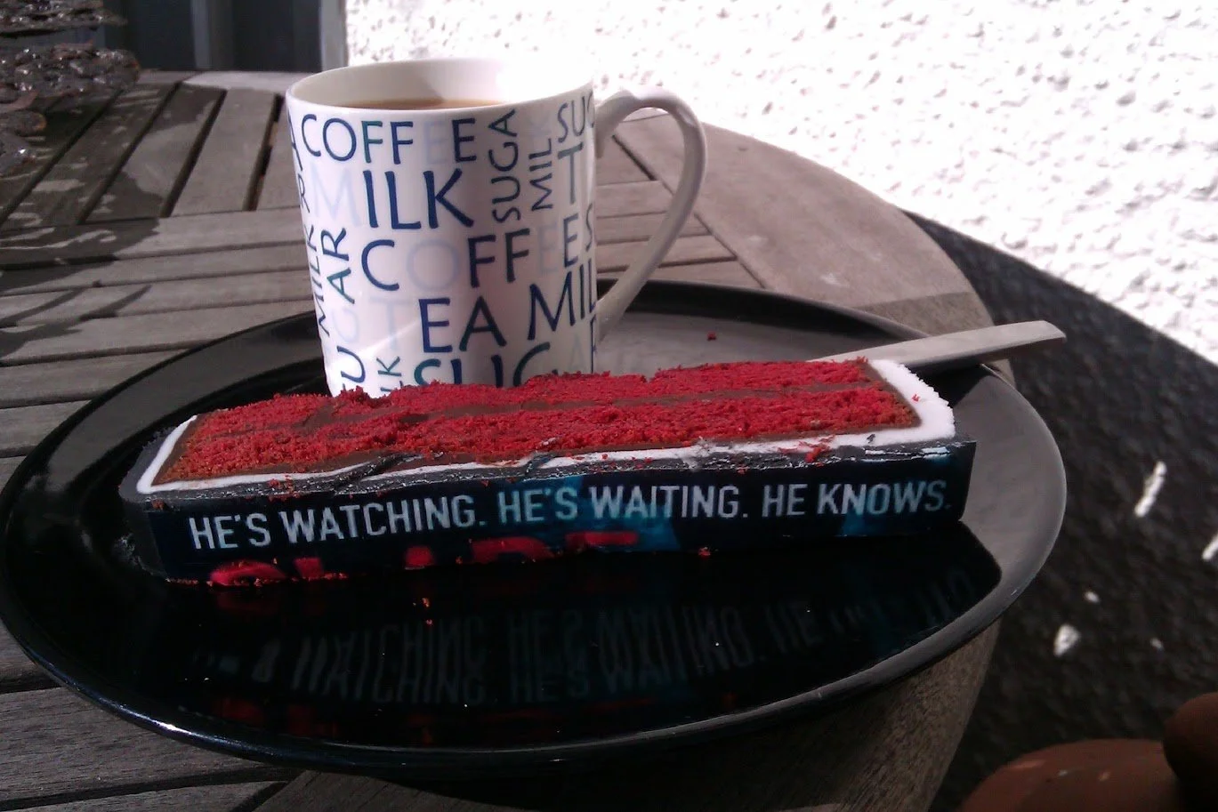 A black tray with a cup of coffee, a cake that looks like a book, on an outdoor wooden table.