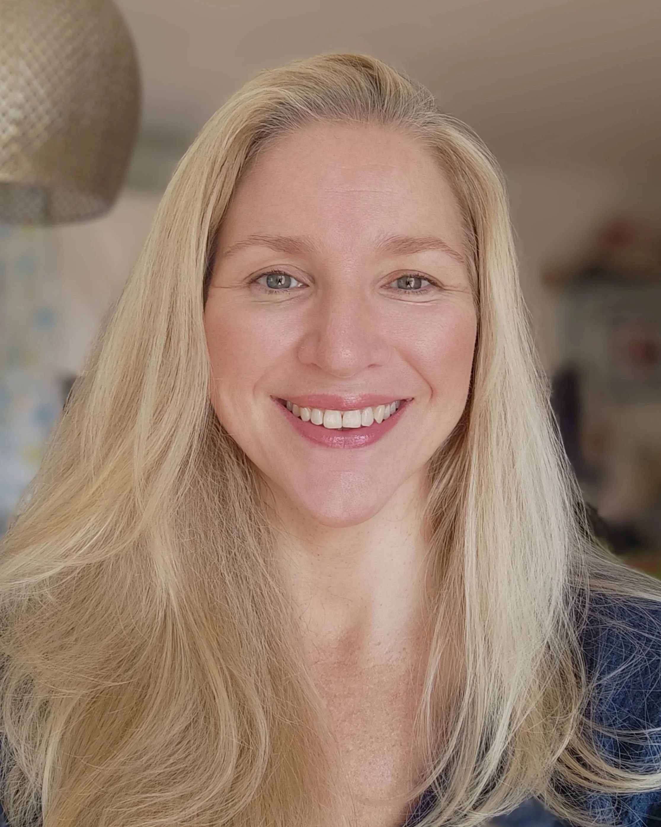 Close-up of a smiling woman with long blonde hair and blue eyes, indoors.