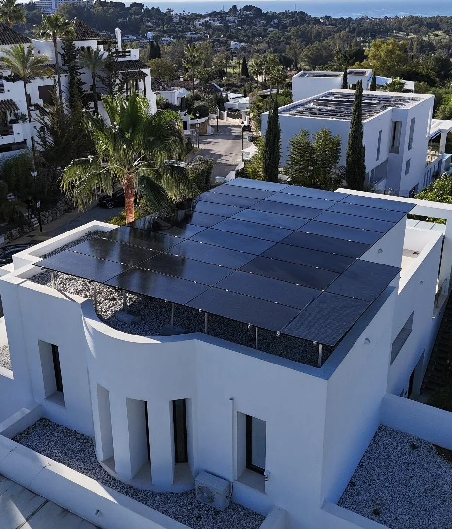 Edificio modernista con paneles solares en el techo, rodeado de árboles y casas blancas en una zona residencial con vista al mar.