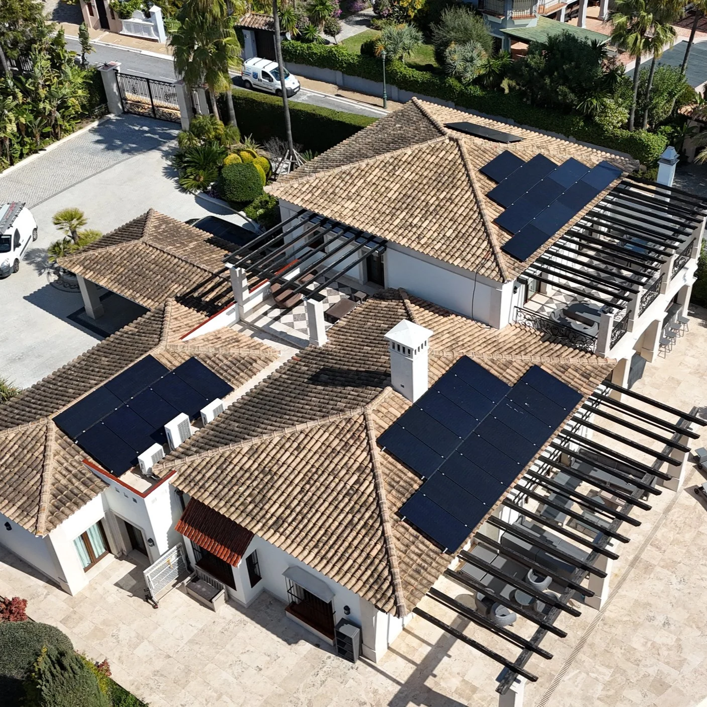 Vista aérea de una casa con tejados de tejas y paneles solares, y áreas de sombra arquitectónicas, rodeada de árboles y una entrada de autopista.