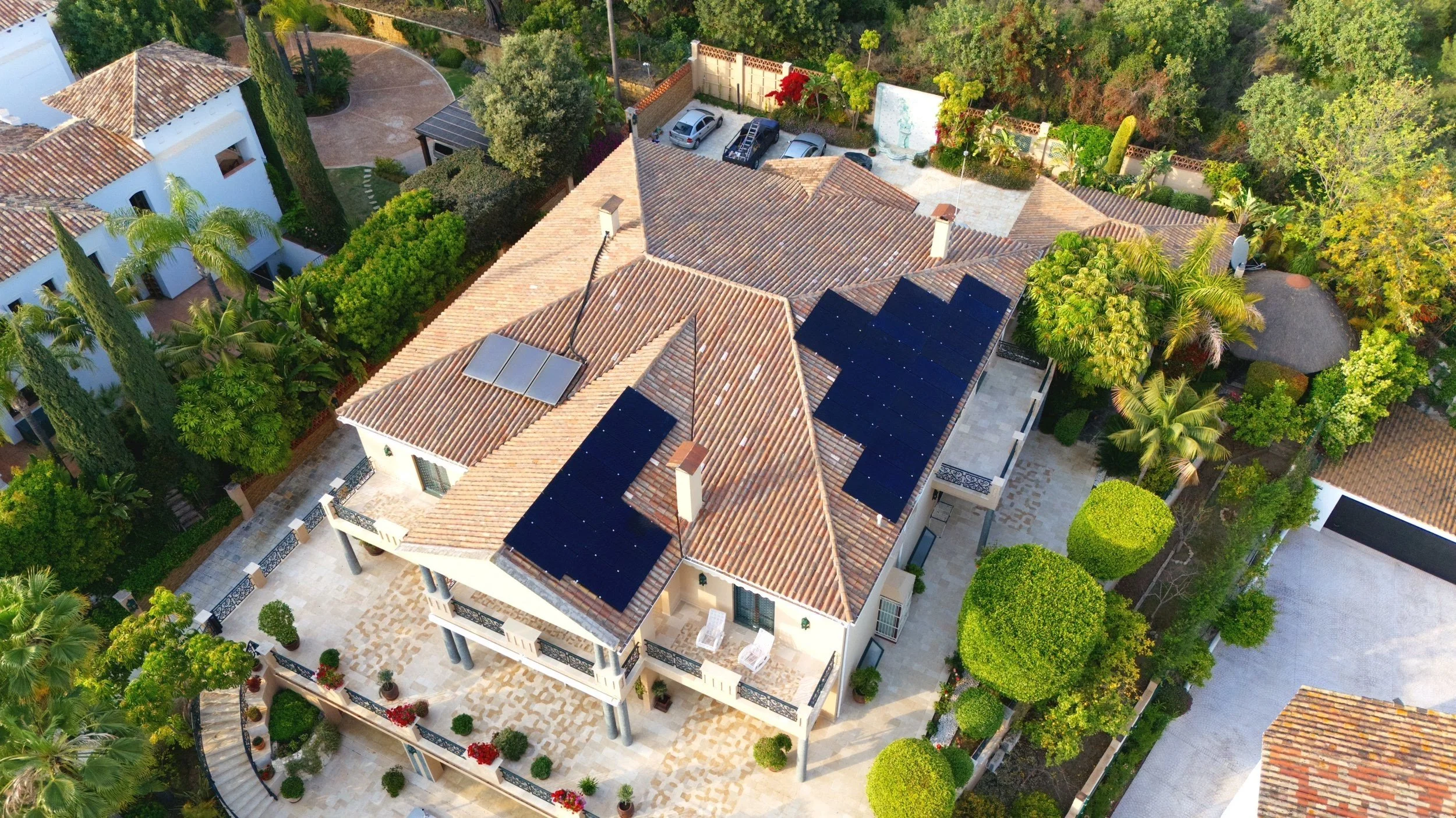 Vista aérea de una casa con tejado de tejas y paneles solares, rodeada de árboles y jardines en un vecindario residencial.