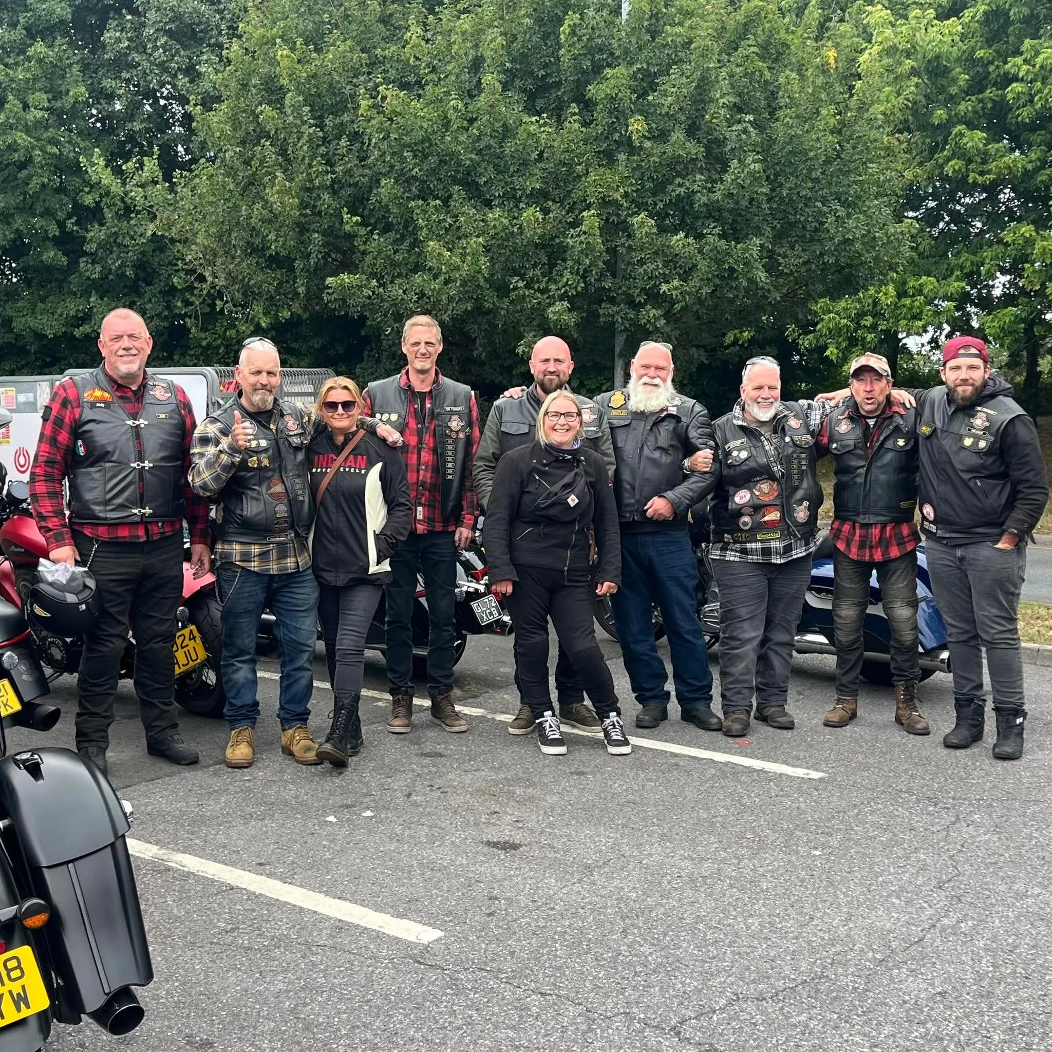 A group of people dressed in Harley Davidson motorcycle club vests standing together outdoors in front of lush green trees.