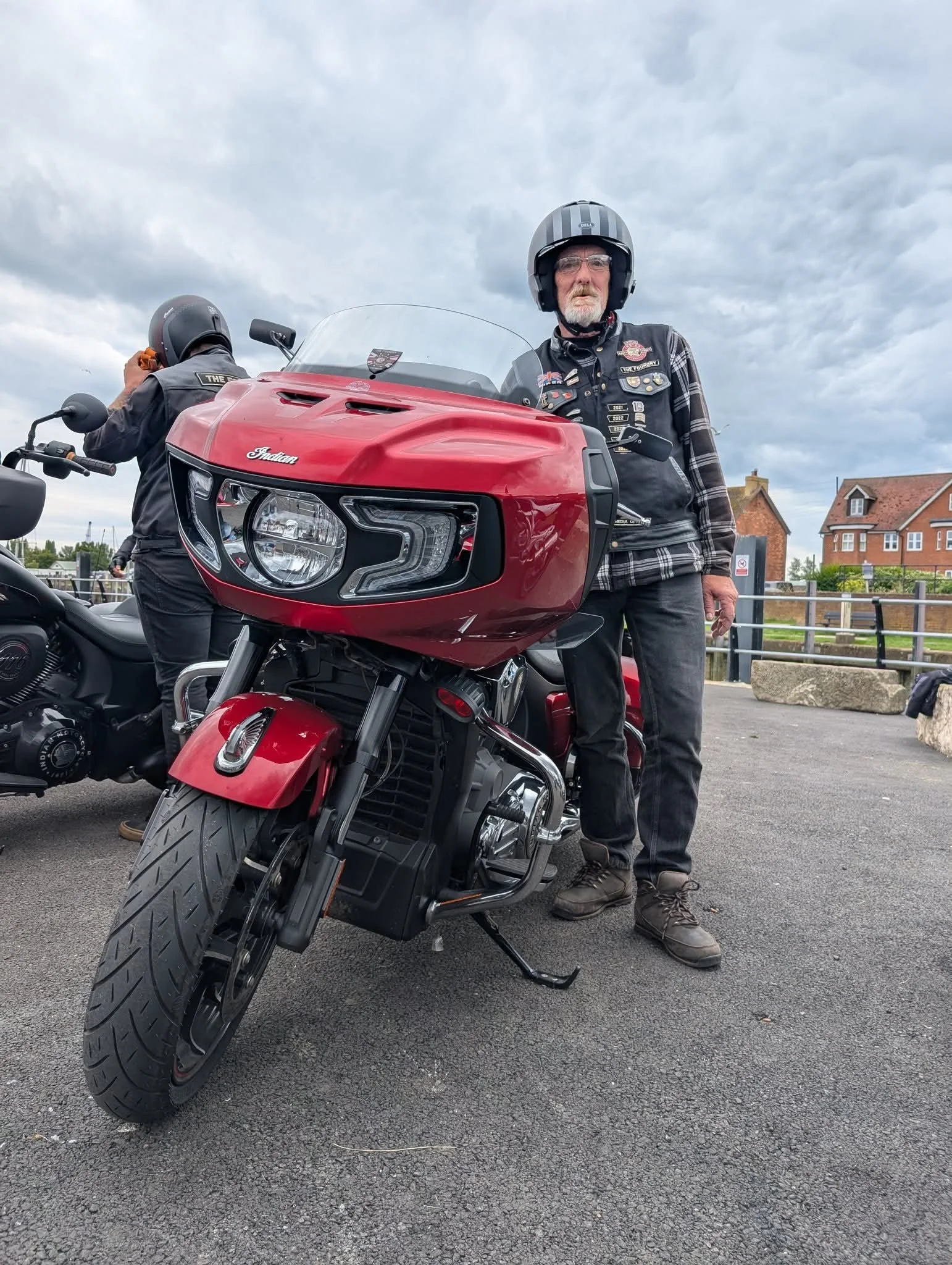 A man standing next to a red Indian motorcycle, wearing a black helmet and a jacket with patches, with a person in the background riding another motorcycle under a cloudy sky in a parking lot with houses nearby.