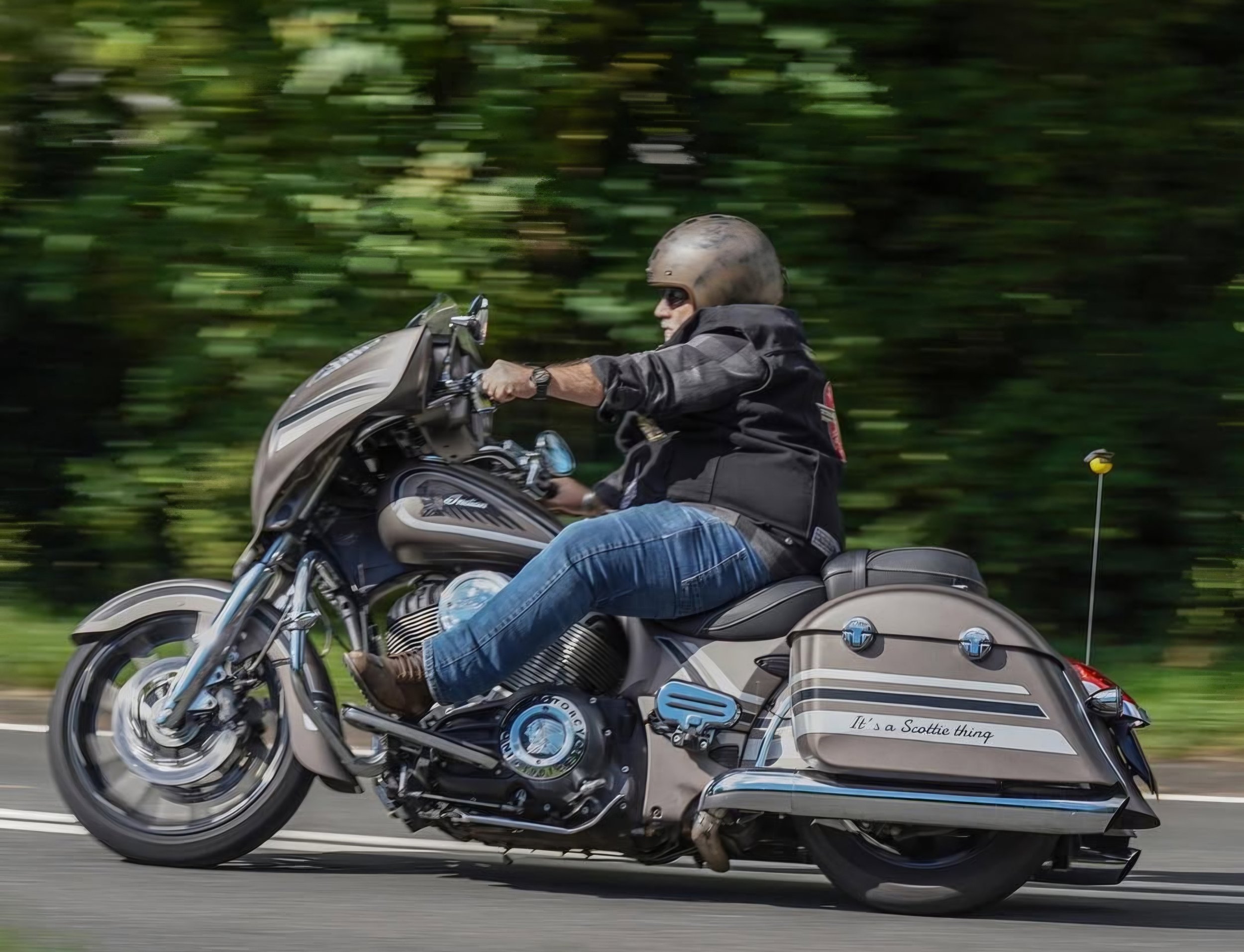 A man riding a motorcycle on a road surrounded by green trees, wearing a helmet and sunglasses, with a leather jacket and jeans.