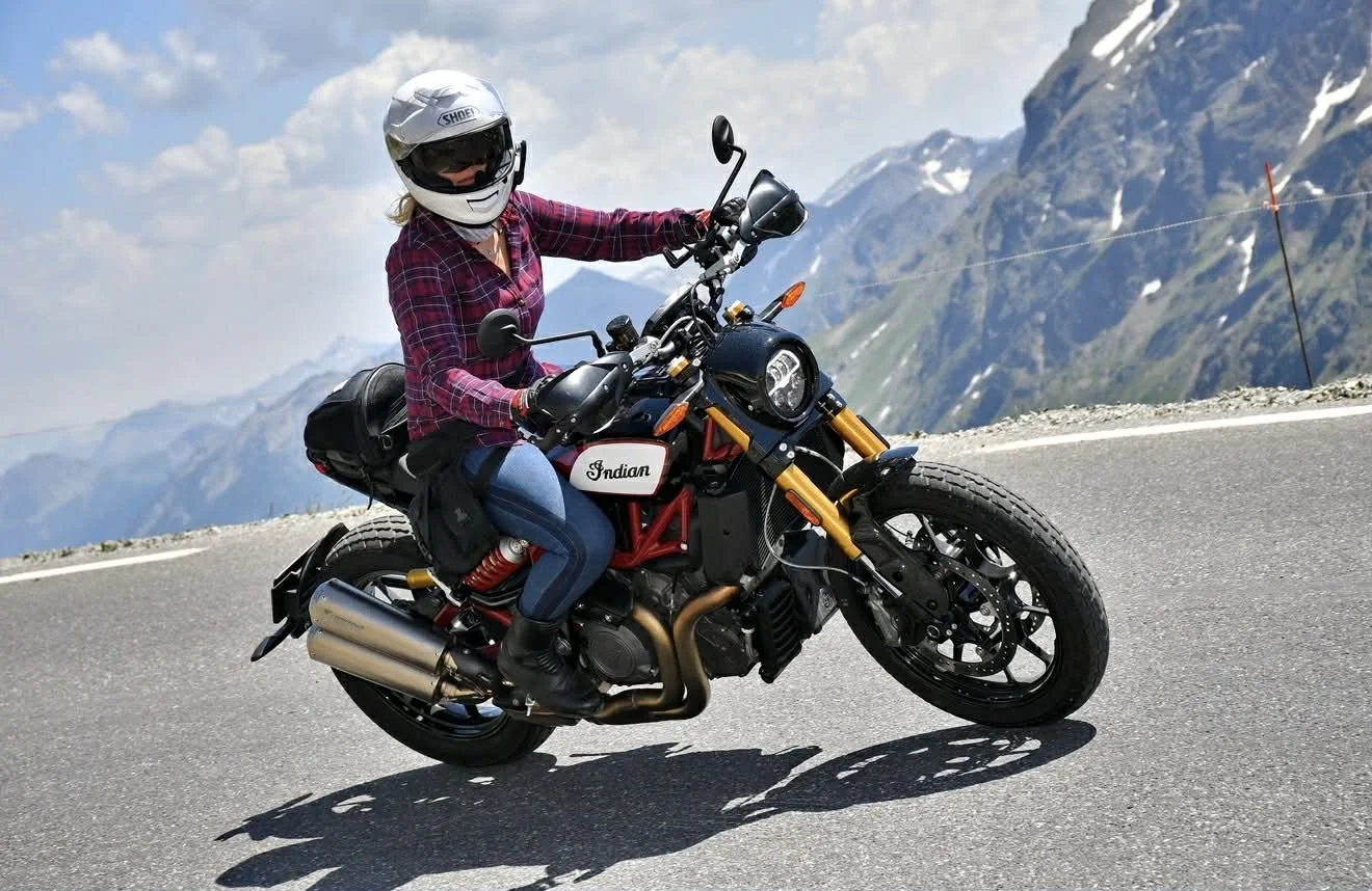 A woman riding a black and red Indian motorcycle wearing a white helmet, sunglasses, a red plaid shirt, and jeans on a mountain road with snow-capped peaks in the background.