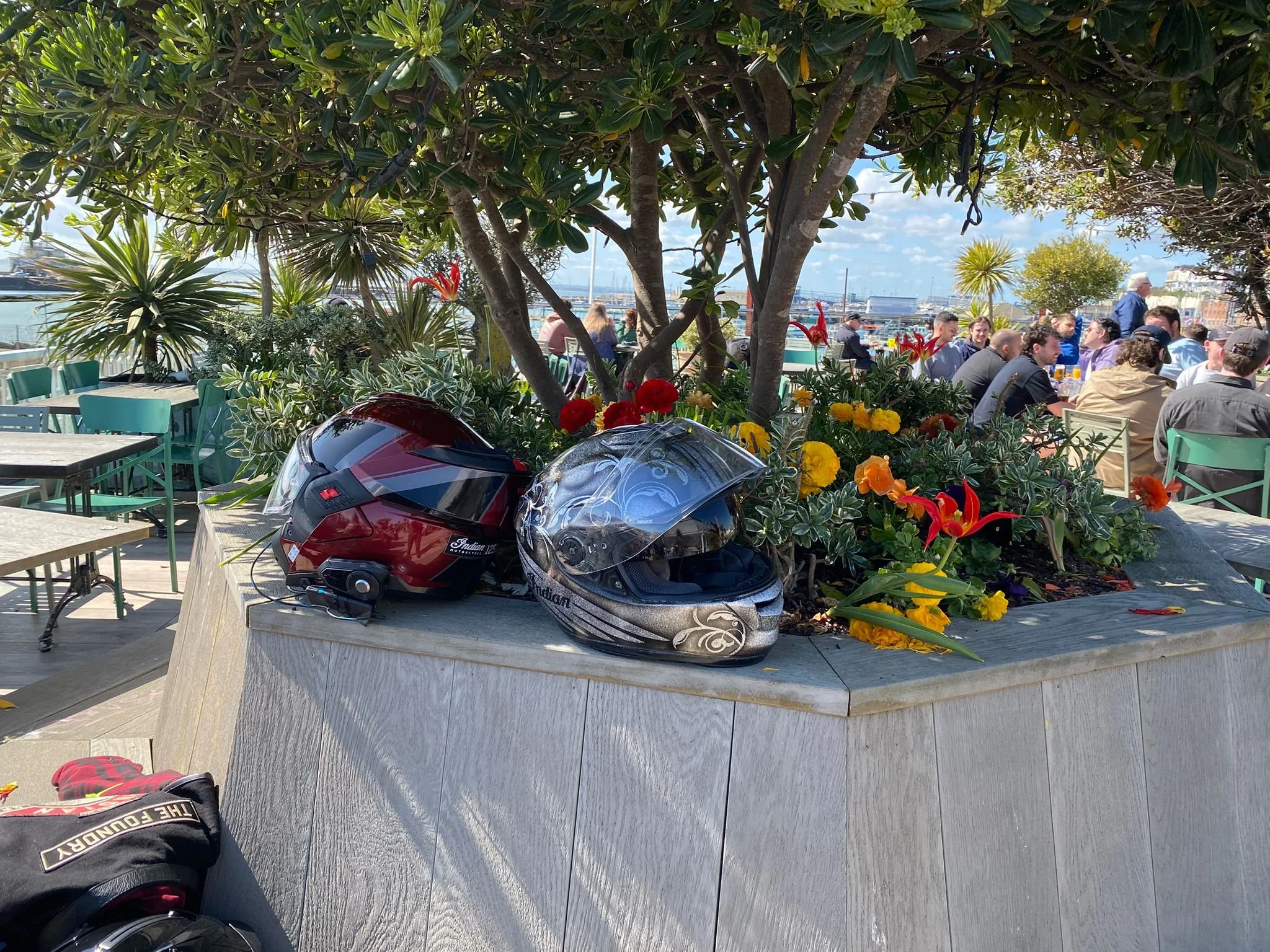 View of a seaside restaurant with outdoor seating, lush plants, colorful flowers, and a marina in the background under a cloudy sky; motorcycle helmets are placed on a table in the foreground.